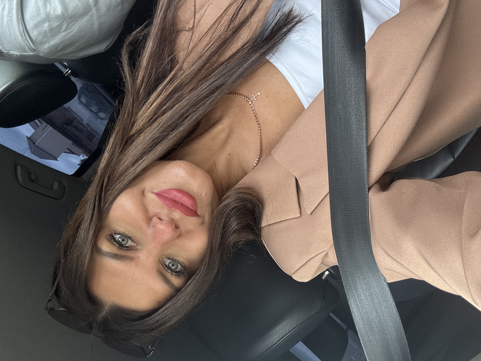 In the city of Chaikovsky, Russia, a woman is captured in a selfie taken from inside a vehicle. She is seated in the driver's seat, with the seatbelt fastened across her. The woman has long, dark brown hair, striking green eyes, and is wearing red lipstick. She is dressed in a light brown blazer over a white top and a delicate rose gold necklace with a small cross pendant. The background reveals the interior of the car, including a black leather seat and a window reflecting buildings and a street scene, suggesting an urban environment.