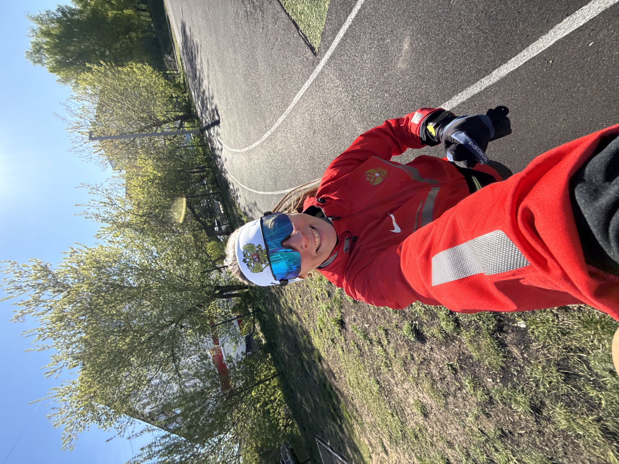 A medium-angle shot depicts a smiling person outdoors, likely an athlete. The person is wearing a red jacket, black gloves, and reflective sunglasses, and a hat with a Russian coat of arms. They are standing on an asphalt track with white lines, possibly a running or cycling track. To the left of the track are trees and glimpses of buildings. The location appears to be a sports facility or park in the city of Chaikovsky, Russia, given the presence of the Russian coat of arms and the sports setting.