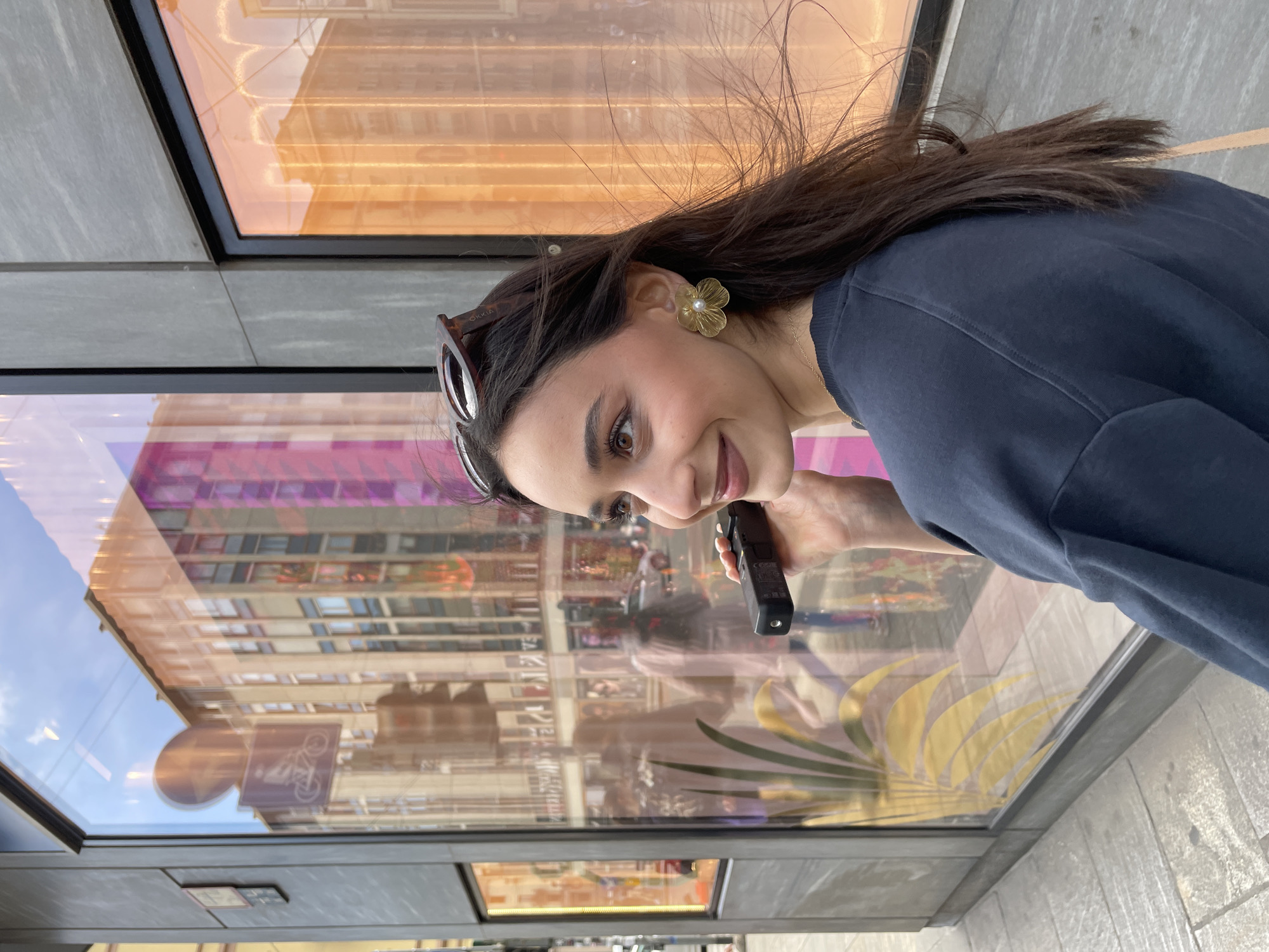 A woman with long dark hair and a navy blue sweatshirt stands in front of a shop window, holding a black object. The building reflected in the shop window has a bicycle sign and appears to be in Milan, Italy, based on visual characteristics. The woman wears a flower-shaped earring and sunglasses are perched on her head. She is looking toward the camera with a slight smile.