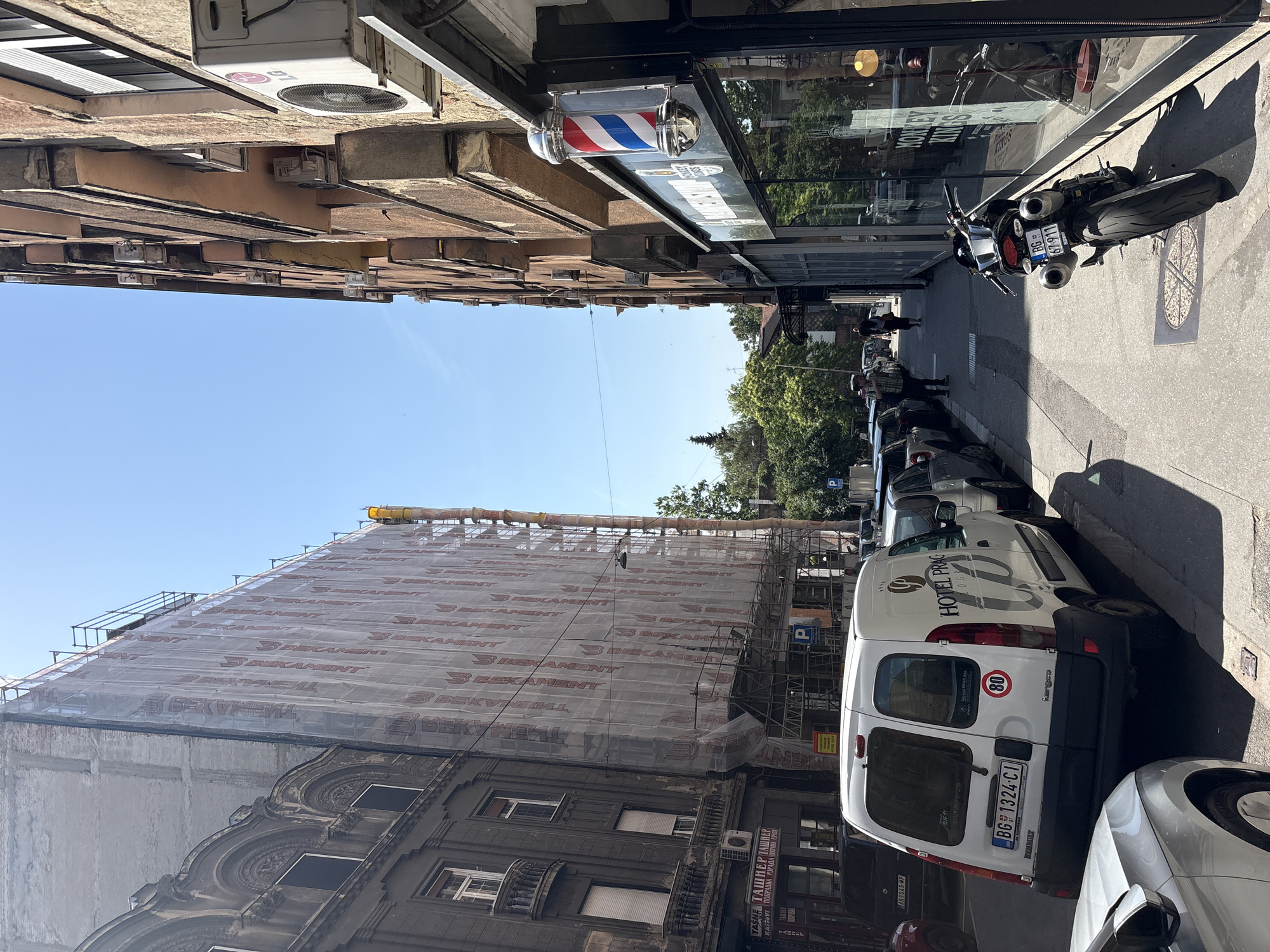 The image captures a street scene in Belgrade, Serbia.
The view looks down a narrow street lined with buildings on both sides. The building on the left is under construction, covered in scaffolding and protective netting labeled "BEKAMENT." The building is dark gray, with ornate architectural details.
On the right, buildings are closer, constructed of tan-colored brick with air conditioning units visible. A barber shop is identifiable by the classic red, white, and blue barber pole hanging outside.
Along the right side of the street, cars are parked. A white van with the logo "HOTEL PRAG" is prominently displayed, and the license plate reads "BG 1324-CI," indicating it is registered in Belgrade. A motorcycle is parked further down the street. Several pedestrians are also visible in the distance, walking along the sidewalk.
The sky above is clear and blue.