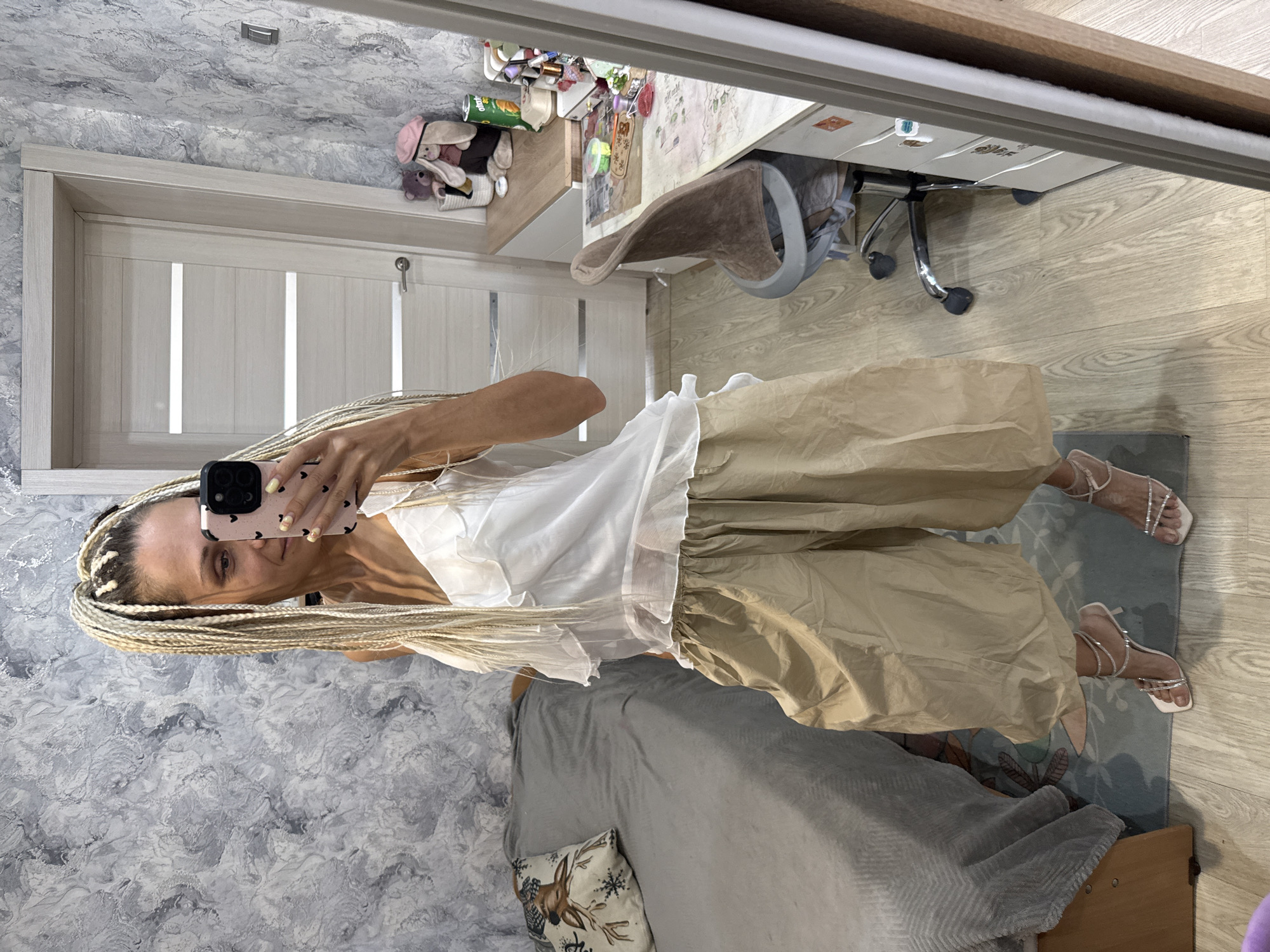 A woman with blonde braided hair is standing in a room and taking a selfie in a mirror. She is wearing a white ruffled camisole and beige, voluminous, wide-leg pants. She is also wearing strappy, jeweled, high-heeled sandals. The room appears to be a bedroom with a grey bed covered with a grey duvet and a pillow with a deer print on it. To the right of the bed is a desk with a chair and various items on it, including a stuffed rabbit and a can. The walls are textured grey. The flooring is light-colored wood. The scene is consistent with an indoor residential environment, possibly in Rusanovo, Russia, given the context provided.