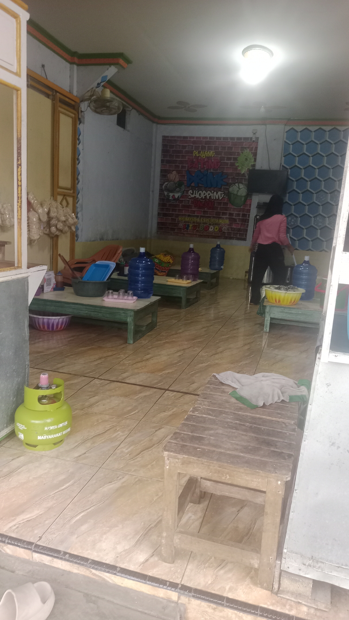 The image depicts the interior of a food stall, likely in Tonjong, Indonesia, as indicated by the text on the gas cylinder. The stall features a tiled floor and a ceiling with a light fixture and a fan. In the background, a brick-patterned mural displays colorful text related to food and drinks, including "PLAYING," "DRINK," and "SHOPPING," along with illustrations of food items.

Several low tables with green wooden frames are set up, some holding large water bottles, plastic containers, and glasses. A woman in a pink shirt and dark pants is visible, standing near a table with a yellow basin. A green gas cylinder is positioned on the floor near the entrance, with Indonesian text indicating it is "ONLY FOR COMMUNITY." A wooden bench with a piece of cloth on it is also present. The overall scene suggests a casual, possibly bustling, small business environment.