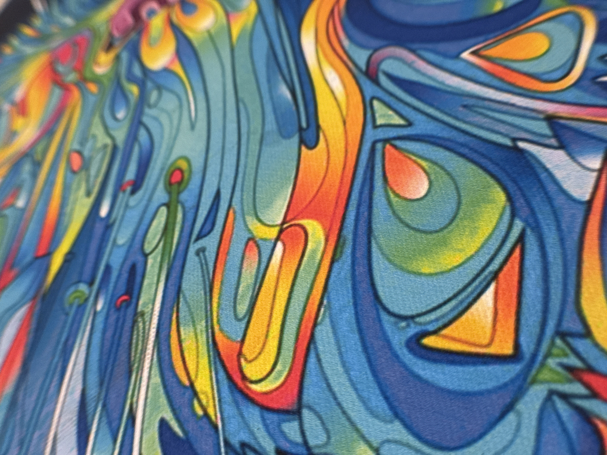 The image shows a close-up view of a colorful, abstract artwork. The dominant colors are shades of blue, yellow, orange, red, green, and white. The composition consists of fluid lines and shapes, creating a sense of movement and depth. Some forms appear to be geometric, like a triangle. The artwork has a textured appearance, possibly indicating it is painted on canvas or another fabric. I cannot find any information related to the location of this work in Novo-Nikol'skoe, Russia.