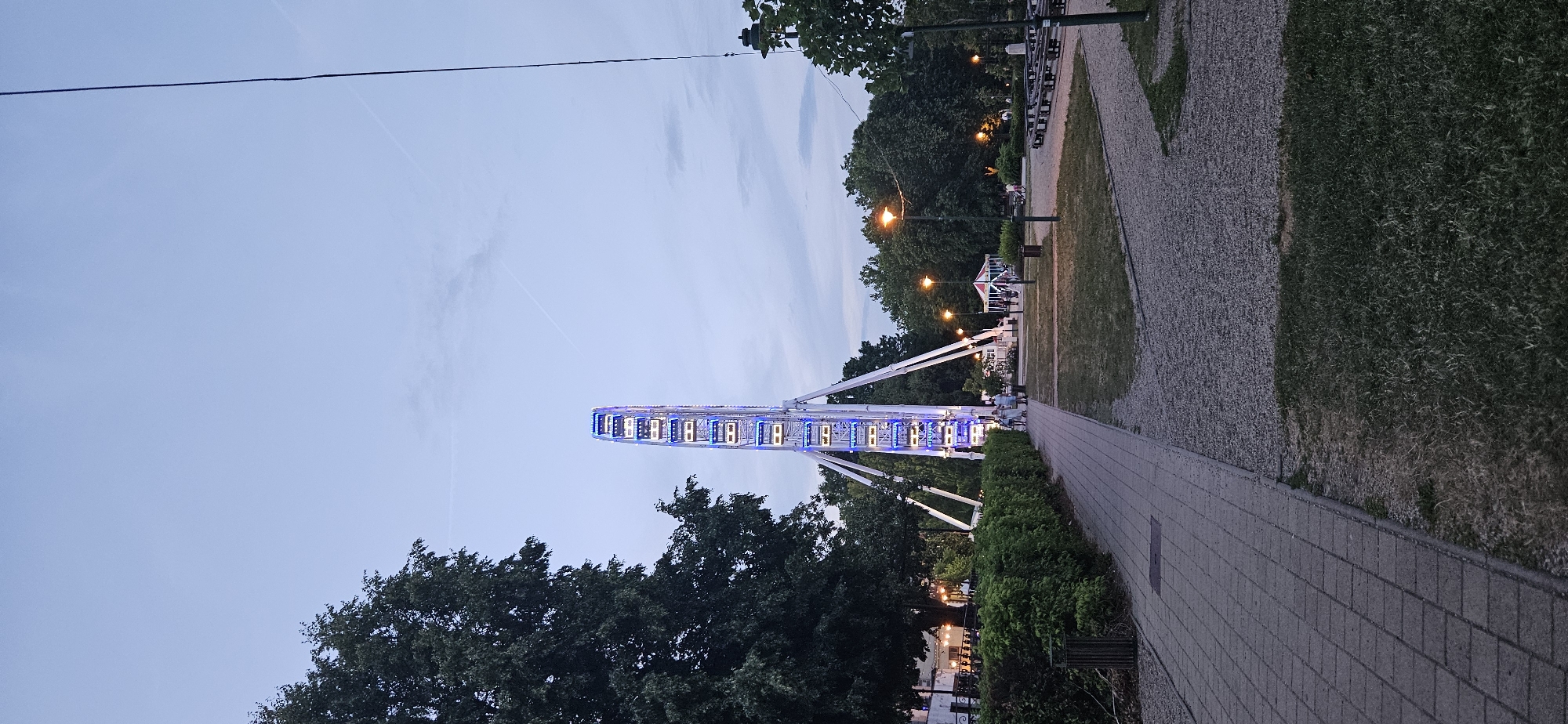 The image depicts a Ferris wheel lit with blue lights, positioned in an amusement park or fairground area in Keszthely, Hungary. The Ferris wheel is the central and dominant feature, standing tall against a pale blue, twilight sky with faint contrails. In the foreground, a paved walkway made of interlocking bricks runs from the left to the center of the image, bordered by a dense, green hedge on the left and a gravel path on the right. Several trees with dark green foliage are visible, one large tree on the far left and more in the background. Park benches are arranged in clusters on the right side of the image. Various park lights are illuminated, casting a warm glow on the surroundings. A small, conical structure, possibly a food stall or a tent, is visible in the middle ground. A few people are seen walking on the path near the hedge and in the background of the park area. The overall atmosphere suggests an evening setting at an outdoor recreational space.