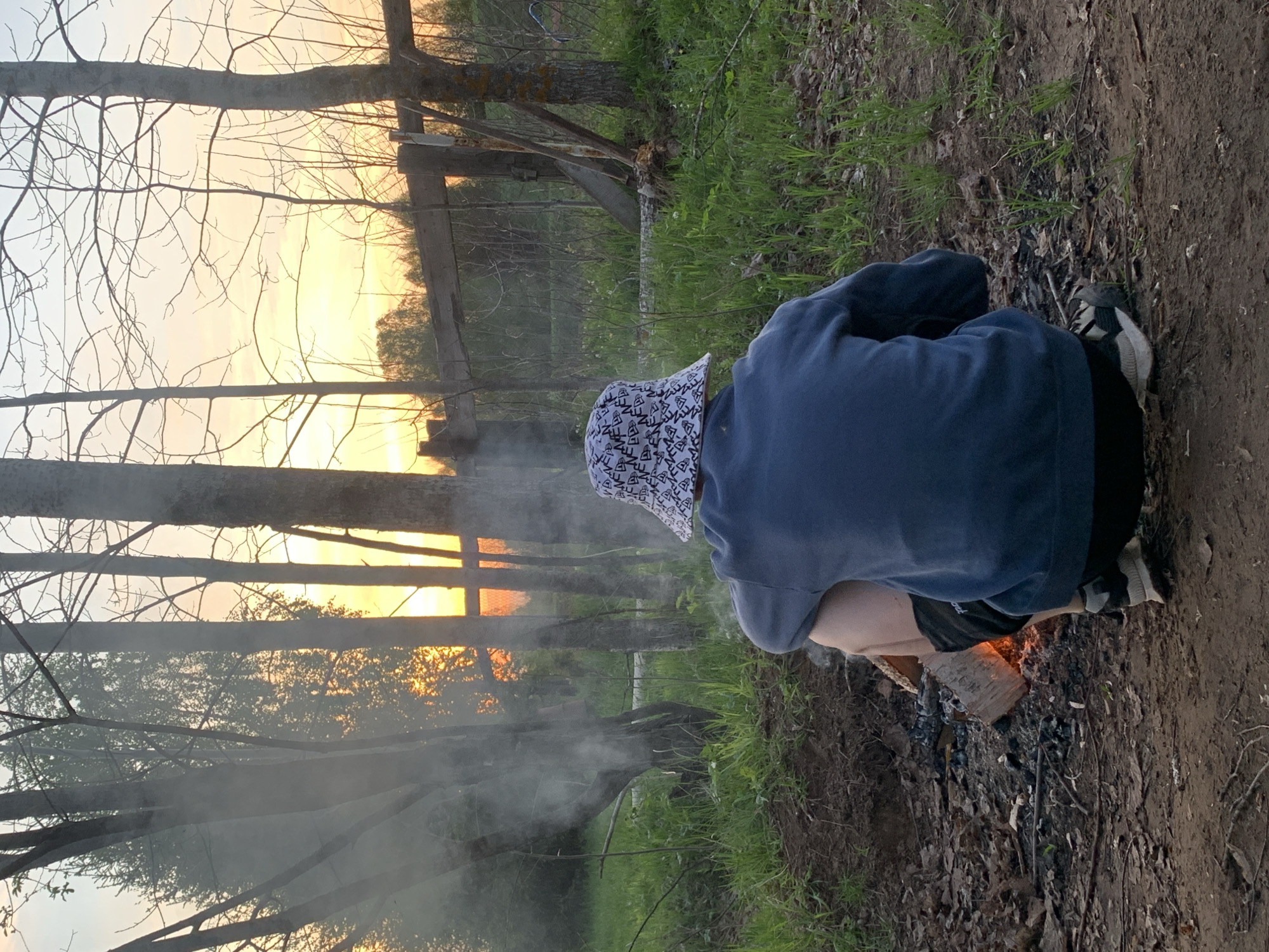 The image depicts an individual crouched near a small fire in a wooded area. The person is wearing a blue long-sleeved shirt, black shorts, patterned bucket hat, and black and white shoes. A burning log is visible amidst charred wood and ashes. Smoke rises from the fire. The background reveals trees, foliage, and a fence. The sky, visible through the trees, suggests either sunrise or sunset. The location is likely Zasechnyi, Russia, based on the presence of tree species commonly found in that region.