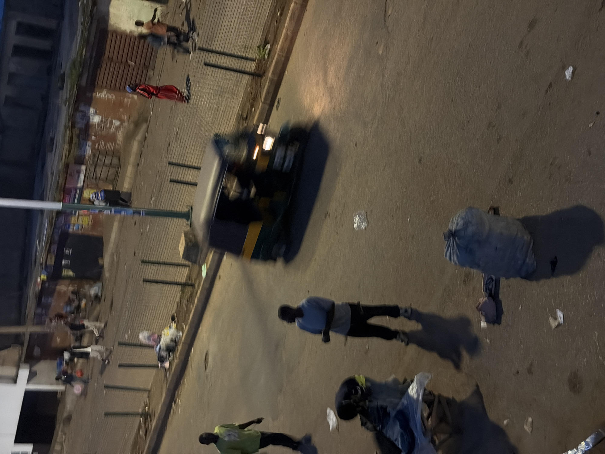 In Bukuru, Nigeria, an outdoor street scene is depicted with a focus on the ground level, capturing a dimly lit, urban environment. Several individuals are present. In the foreground, a person in a light blue t-shirt and dark pants stands, looking towards the left. To their left and slightly in front, another person is bent over, appearing to interact with some items on the ground. Further to the left, a person in a neon green t-shirt and dark pants walks away from the camera. In the mid-ground, a three-wheeled auto-rickshaw is moving towards the right side of the frame, its headlights illuminated. In the background, more individuals are visible walking on the street, some carrying objects. The street is paved, with debris and discarded items scattered across it. A low fence runs along the right side of the street, separating it from an area with buildings. The overall impression is one of a bustling, albeit somewhat disorganized, urban streetscape.