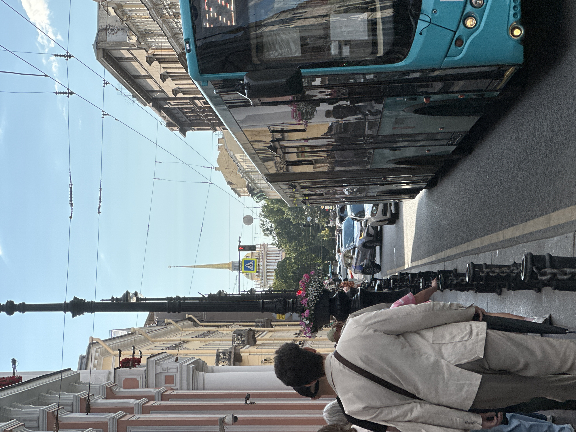 The image depicts a street scene in Saint Petersburg, Russia, likely near the Admiralty building, as indicated by the prominent spire in the background. A teal-colored bus occupies the right side of the frame, facing away from the viewer. In the foreground, a person wearing a light-colored jacket and pants walks away from the camera, carrying an umbrella. Several other pedestrians are visible behind this individual, some partially obscured. The street is lined with ornate buildings on the left, featuring classical architectural elements such as columns and balconies adorned with flowers. Overhead, a network of trolleybus wires crisscrosses the sky. Traffic, including cars and the bus, is present on the asphalt road. The sky is a clear blue with a few scattered clouds, and sunlight illuminates the scene, casting shadows.