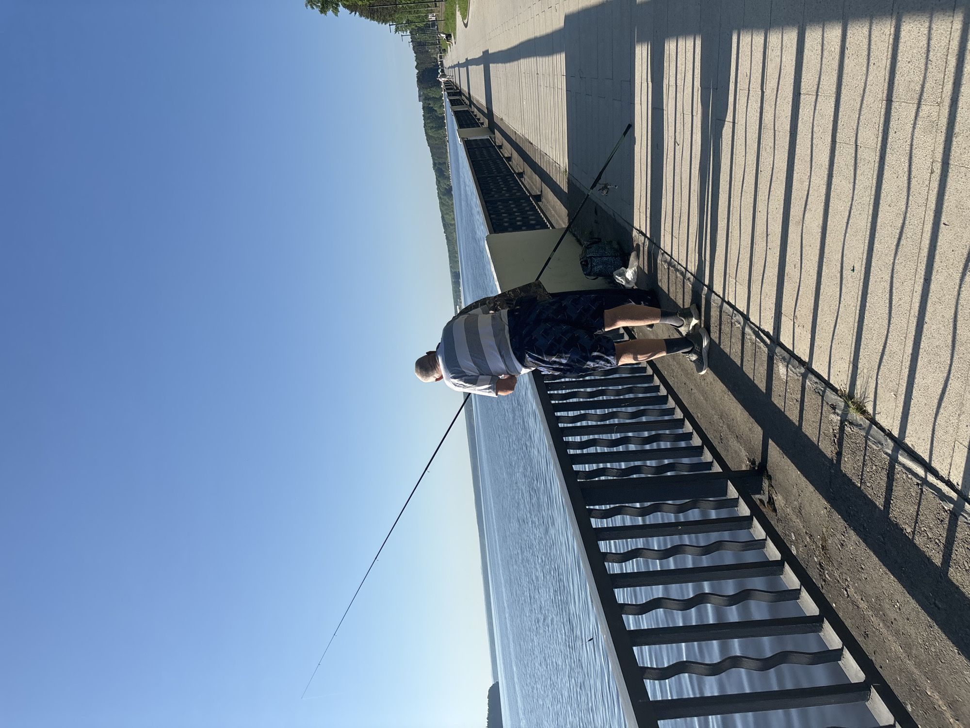 The image depicts a man fishing from a pier in Chaikovsky, Russia. He is wearing a striped shirt and dark patterned shorts, with sneakers on his feet. He is positioned on the pier with a fishing rod extended over the water. Another fishing rod leans against the pier wall near a bag. The pier has a distinct black metal railing with a wave-like pattern. The walkway of the pier is made of light-colored stone or concrete, with the shadows of railings visible. The water is calm and reflects the clear sky. Along the pier, trees are visible in the distance.