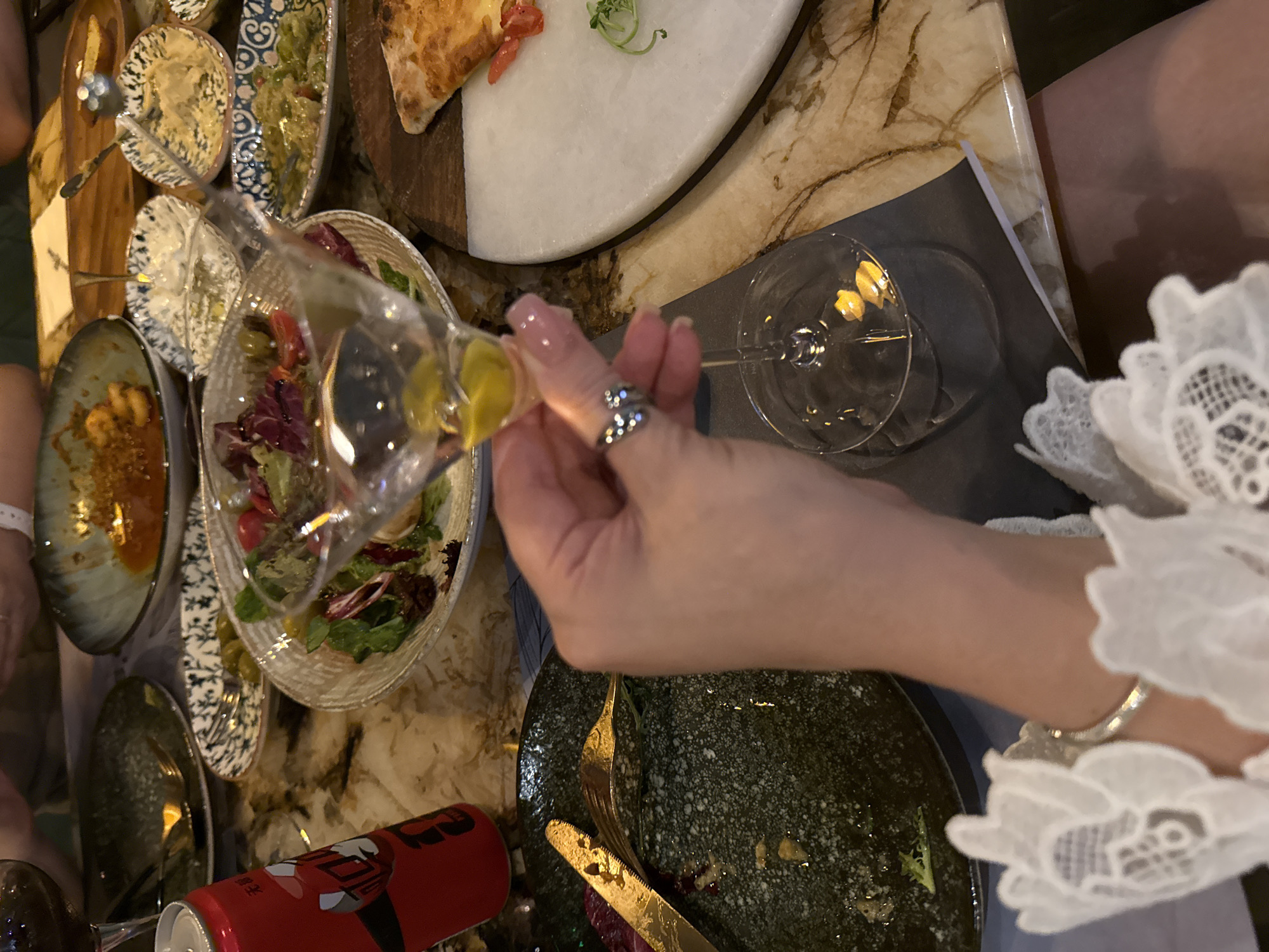 The image depicts a close-up, overhead view of a table at a restaurant. The table is covered in a marble-patterned surface. In the foreground, a person's hand is holding a martini glass with yellow liquid and some ice. The person is wearing a ring on their index finger and a lace-trimmed white garment on their wrist. Next to the hand is a dark, speckled plate with remnants of food and a gold fork and knife. To the left of this plate is a red can with Chinese characters, likely a beverage.
Further back on the table, several dishes are visible. These include a salad in a white bowl, small bowls with dips or appetizers, and a slice of pizza on a wooden board. The overall setting appears to be a meal being shared, with various plates of food and drinks spread out. The lighting suggests an indoor environment, possibly a restaurant in the city of Yiwu, China, given the presence of the red beverage can with Chinese writing. The focus is on the dining experience, with the hand and martini glass being the central element in the immediate foreground.
