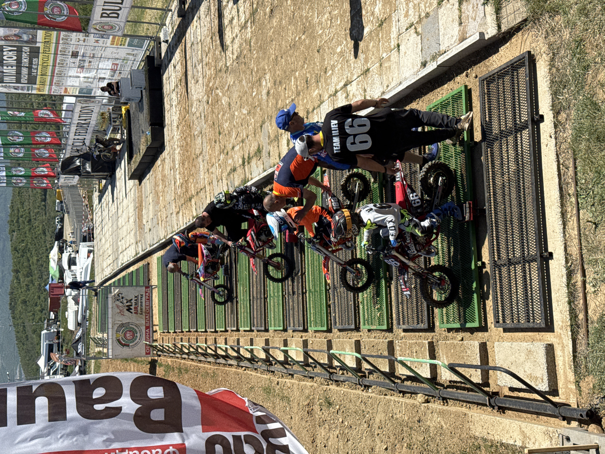 The image shows a motocross starting line with several racers on their bikes, accompanied by their crew members. The racers are positioned on a metal starting gate system. A person wearing a black shirt with the number "66" and the name "TEAM ILIEV" is standing next to one of the racers. Various banners and signs are visible in the background, including one that says "IN MEMORY КРАСИМИР БОЖУРСКИ 16 АПРИЛ 2026 BULGARIAN MOTORCYCLE FEDERATION" and another that includes "MX AIRMAX БЛАГОЕВГРАД ОЕ ДИНА БЛАГОЕВГРАД" . Part of another banner or flag with the text "Bau" is also visible in the foreground. The location is in the city of Selishte, Bulgaria.