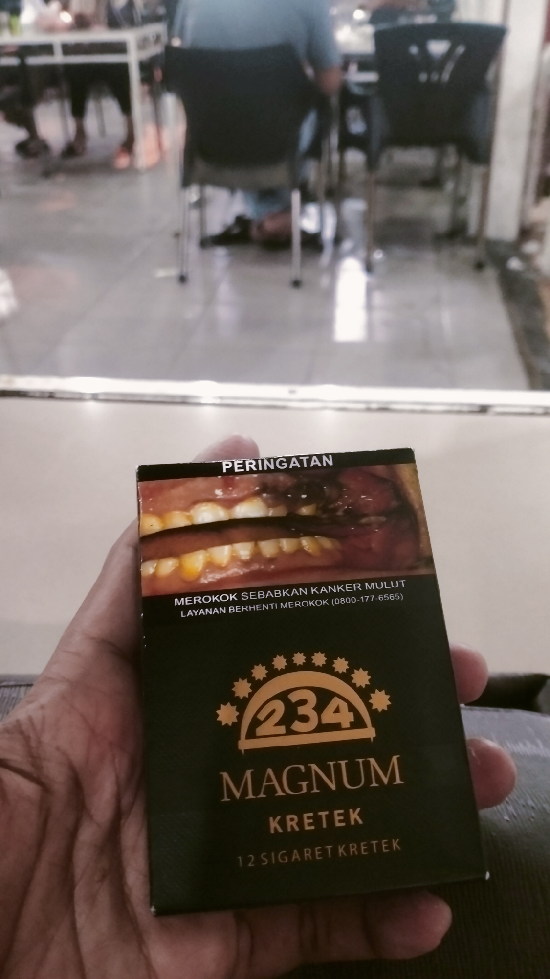 A close-up, eye-level photograph captures a right hand holding a packet of "Magnum Kretek 234" cigarettes in an indoor setting in Lhokseumawe, Indonesia. The hand, with a light brown skin tone, visible veins, and subtle wrinkles, is positioned in the lower center, gripping the dark green/black and gold cigarette pack. The pack's front is in sharp focus, displaying the brand name "MAGNUM KRETEK" with a stylized "234" logo featuring stars, and the text "12 SIGARET KRETEK" (12 clove cigarettes).
Above the brand information is a prominent graphic health warning. The warning text, in Indonesian, reads "PERINGATAN" (WARNING) at the top, followed by "MEROKOK SEBABKAN KANKER MULUT" (SMOKING CAUSES ORAL CANCER) and "LAYANAN BERHENTI MEROKOK (0800-177-6565)" (QUIT SMOKING SERVICE). The graphic depicts a diseased human mouth with discolored, yellowish teeth and a large, dark, ulcerated lesion on the inner cheek or gum, consistent with advanced oral disease.
The background is heavily blurred, indicating a shallow depth of field. It depicts what appears to be an indoor commercial space, likely a cafe, restaurant, or food court. Light-colored tiled flooring is visible, reflecting some ambient light. Indistinct human figures are seated at light-colored tables with dark chairs. One figure in the mid-background wears a light blue top and dark trousers, facing away from the camera. The overall lighting is soft and diffused, characteristic of indoor artificial illumination.
