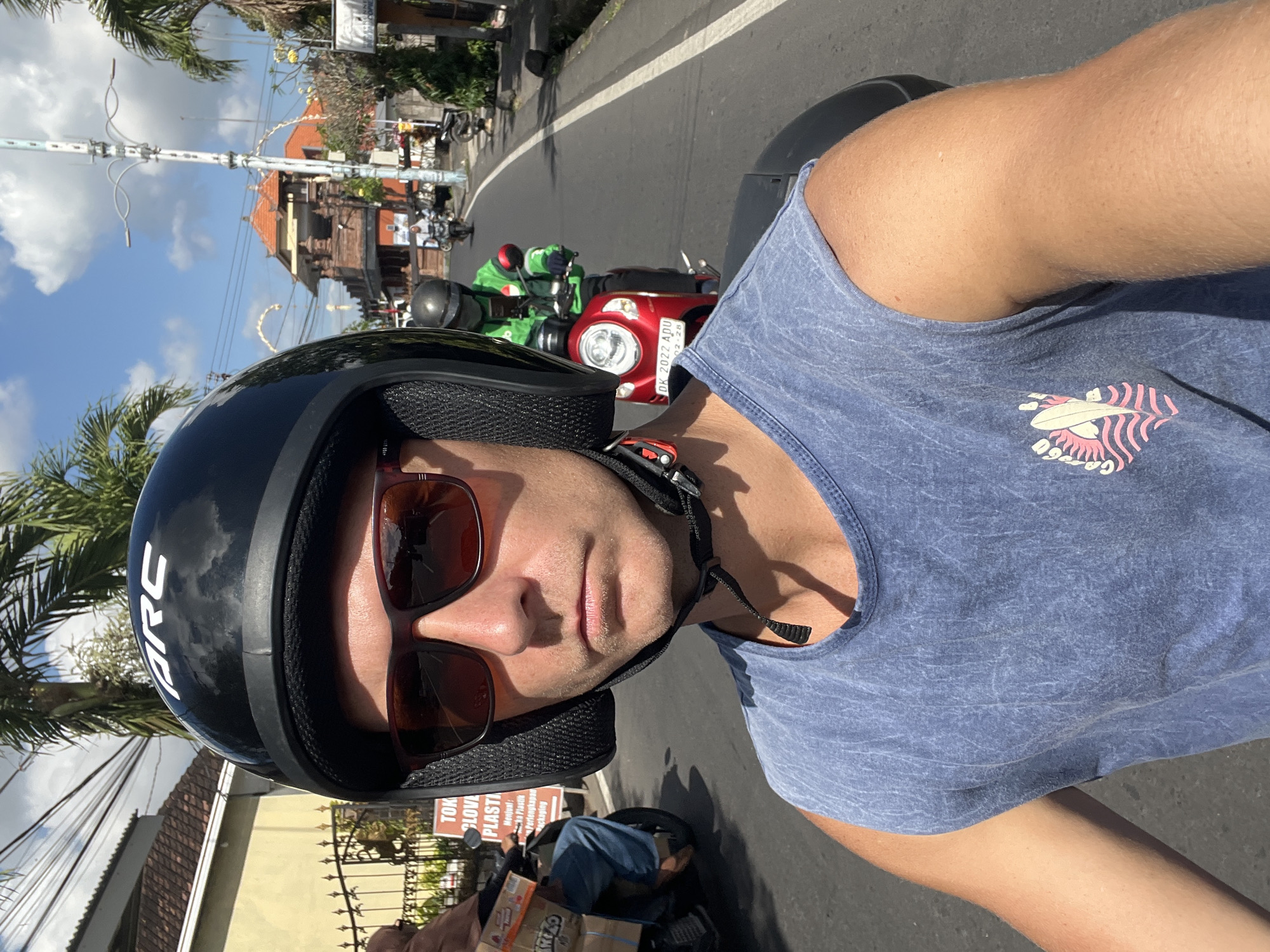 A man wearing a black helmet and sunglasses is riding a scooter on a sunny day. He is wearing a blue sleeveless shirt with a graphic design on it. The scooter has a red body and a license plate that reads "DK 2022 APU". In the background, other people are riding scooters, and there are buildings and palm trees visible. The scene appears to be in a street in North Kuta, Indonesia, characterized by its tropical climate and vibrant local culture.
