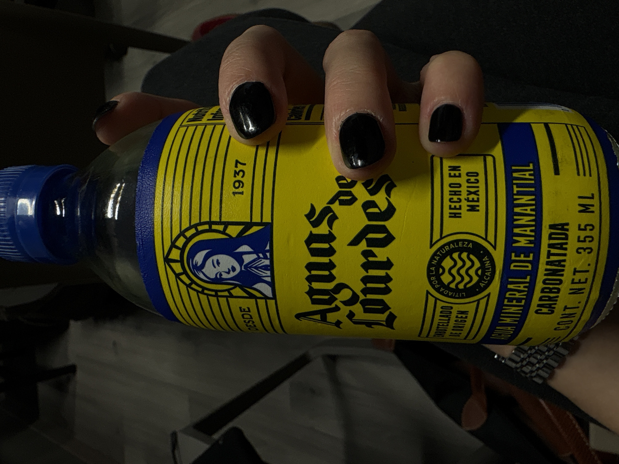 A human hand with polished black fingernails is gripping a bottle of Aguas de Lourdes mineral water. The bottle is yellow with blue trim and a blue cap. The label reads "Aguas de Lourdes" in black lettering, with "Desde 1937" and "Hecho en Mexico" also visible. There is a drawing of a woman on the left side of the label. The text "Agua Mineral de Manantial Carbonatada Cont. Net. 355 ml" is on the right side. The bottle appears to be laying on a surface with dark colors, possibly clothing or a seat.