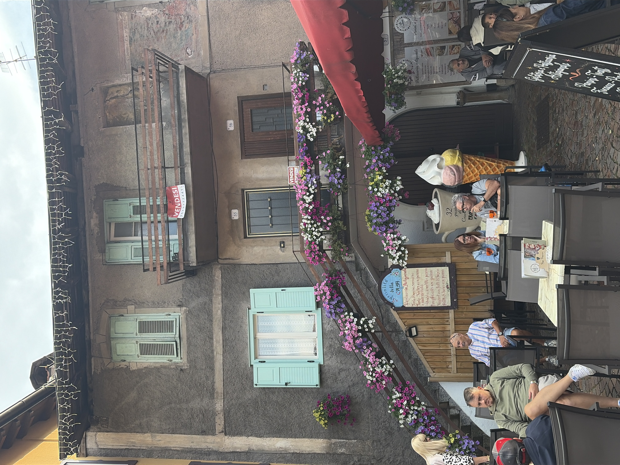 The image shows a building with a gray facade adorned with many colorful flowers cascading down the exterior stairs and balconies. On the upper balcony, a red "VENDESI" (For Sale) sign is displayed. Below, an outdoor seating area with tables and chairs is set up, suggesting a cafe or restaurant. Several people are seated at the tables, some conversing, others drinking. A large, decorative ice cream cone stands near the entrance to the building. The scene is likely located in the pedestrian area of Moena, Italy, a popular tourist town in the Dolomites, given the architectural style and the presence of a gelato shop. The overall atmosphere appears to be casual and relaxed, with a touch of European charm.