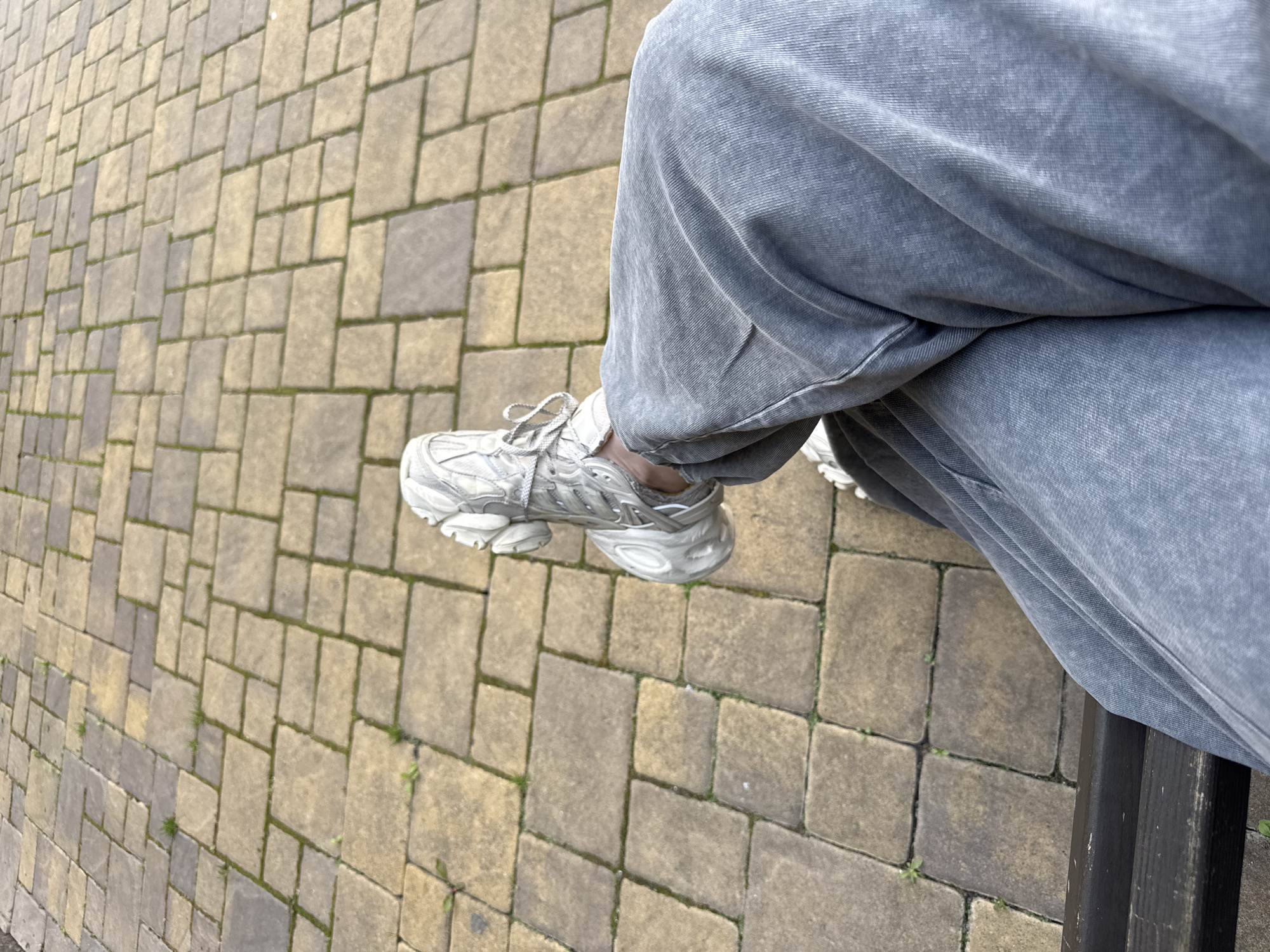 The image captures a close-up, high-angle view of a person's lower body, specifically their legs and feet, while they are seated outdoors. The individual is wearing loose-fitting, faded grey sweatpants or similar comfortable trousers that extend over their ankles. On their feet are white and grey athletic sneakers with a chunky sole and visible laces, typical of a 'dad shoe' or 'ugly sneaker' style.
The person appears to be sitting on a dark-colored bench, a section of which is visible along the right edge of the frame. Their legs are extended forward, with one foot partially off the ground, suggesting a relaxed posture.
The ground beneath and around the person is paved with interlocking concrete pavers. These pavers are a mix of light brown/tan and grey tones, laid in an irregular pattern of rectangular and square shapes. Small patches of green grass or weeds are growing in the crevices between the pavers, indicating an outdoor setting such as a sidewalk, park, or public square. The lighting suggests it is daytime, possibly overcast, given the lack of harsh shadows. The overall scene takes place in Sofiivska Borshchahivka, Ukraine. There is no visible text in the image.