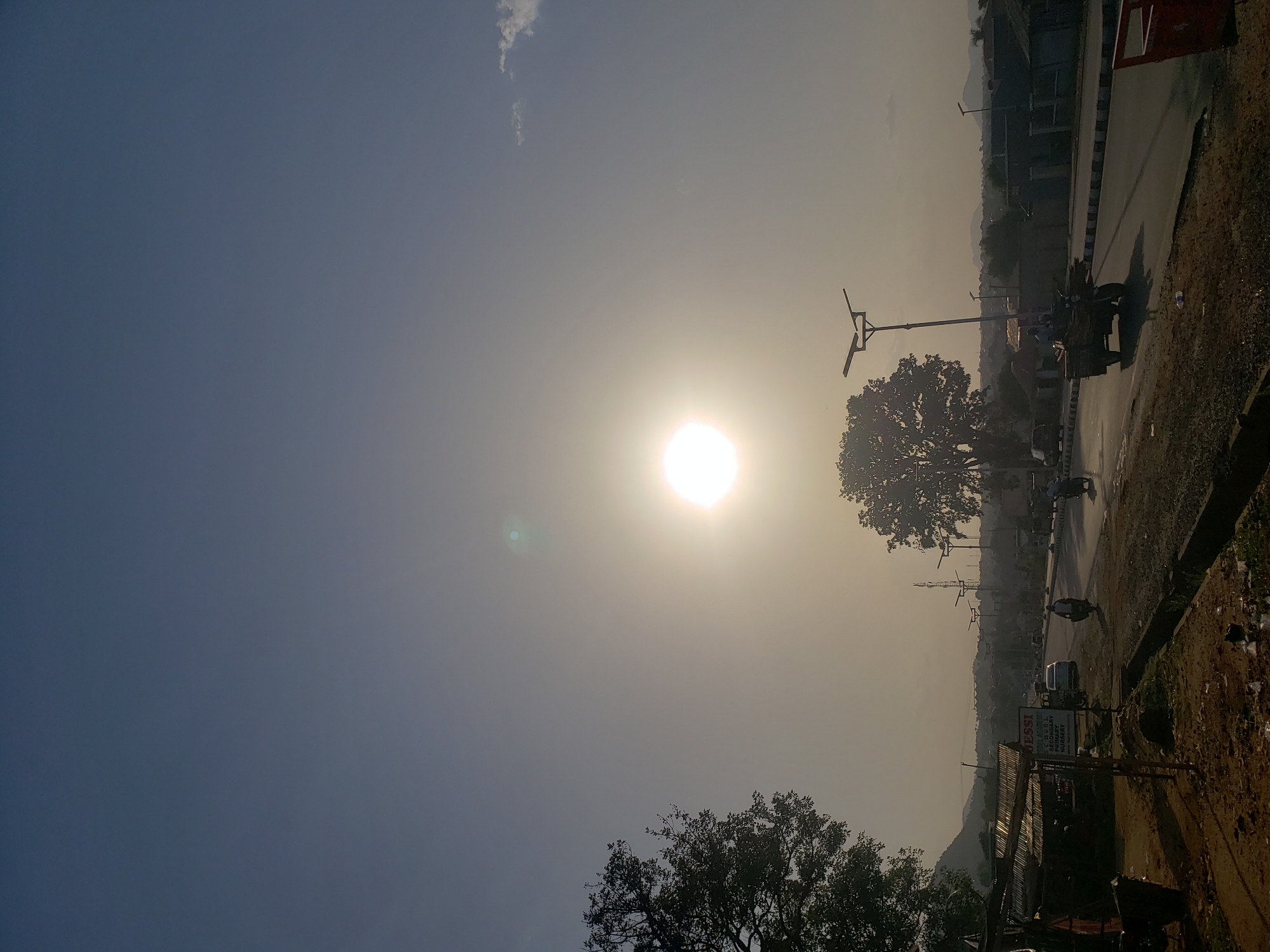 The image depicts a street scene in Adankolo, Nigeria under a bright, hazy sun. A road runs through the frame with a variety of vehicles including cars and motorbikes. Streetlights line the road. Trees are visible on both sides of the road. The sky is hazy. A sign for "Jessi Secondary Nursery" is visible. A mountainous landscape can be seen in the background.