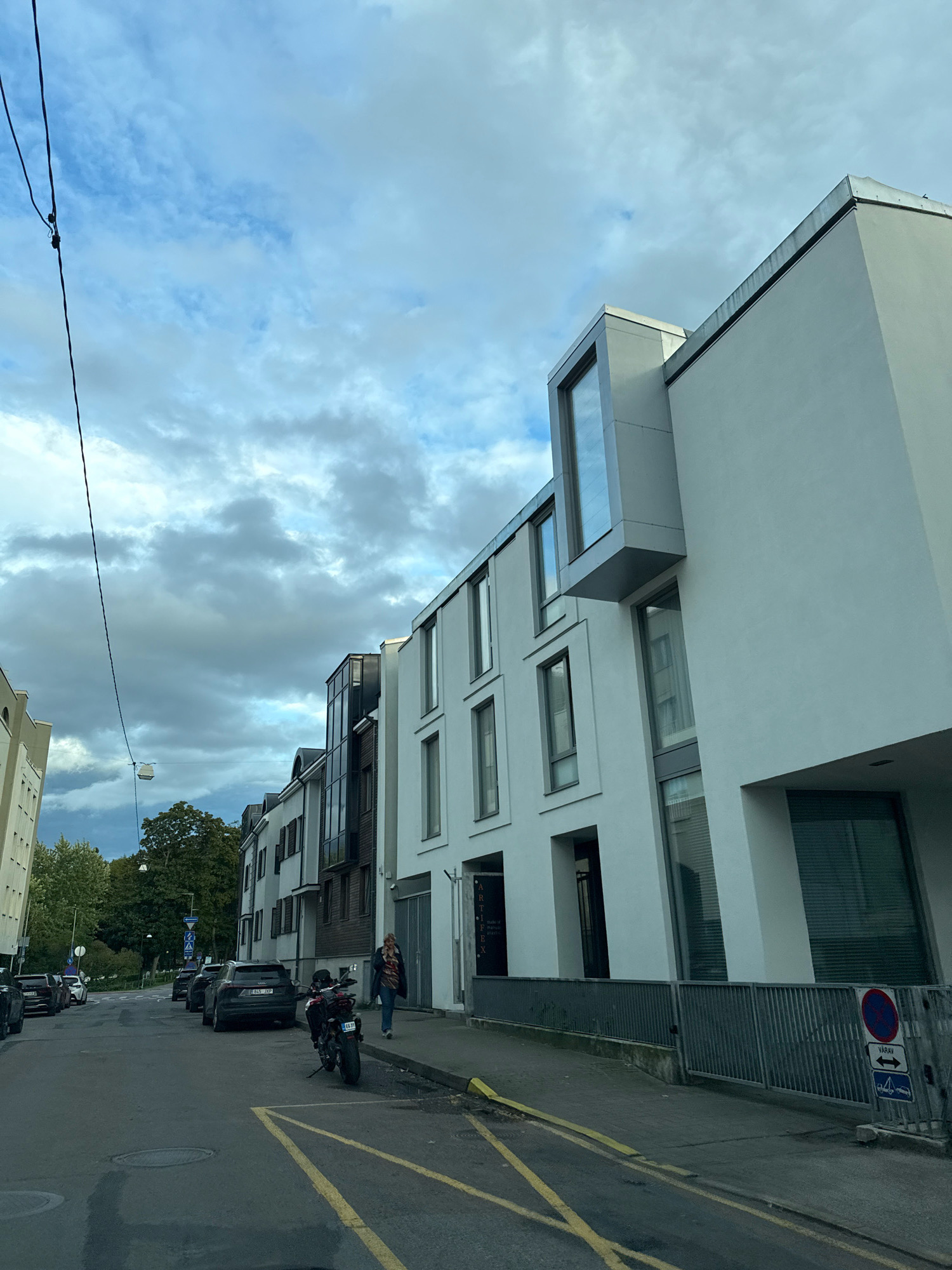 The image shows a street scene in Tallinn, Estonia. On the right side of the street are modern, white buildings with large windows. In the foreground, a motorcycle is parked on the street. A person is walking on the sidewalk towards the buildings. On the left side of the street, there are more parked cars and trees. The sky is cloudy.