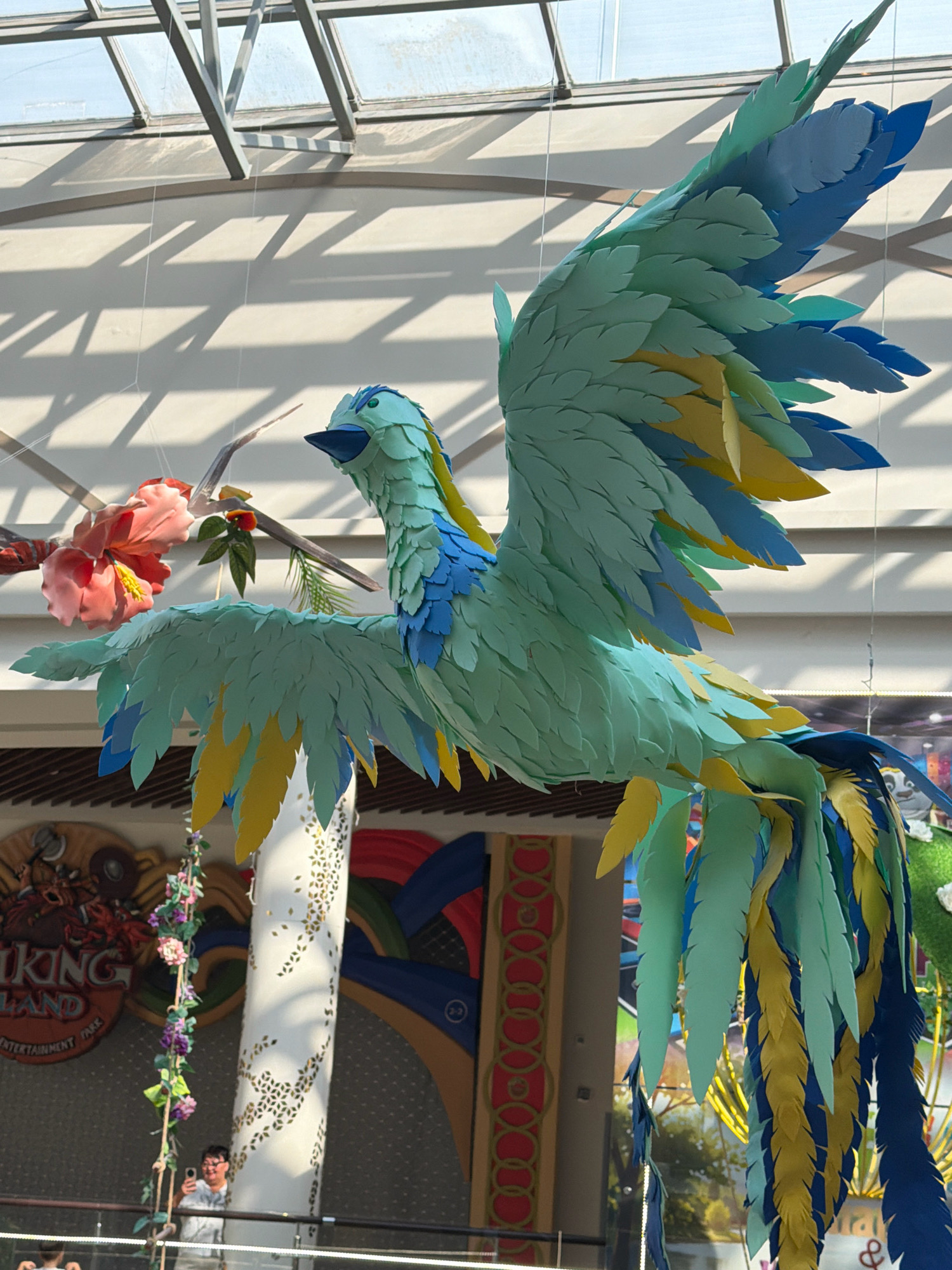 A large, colorful, artificial bird decoration, likely made of paper or foam, hangs suspended indoors. The bird is predominantly teal and light blue, with accents of yellow and darker blue feathers on its wings and tail. It has a distinctive beak and eyes. The background reveals an indoor setting with a glass ceiling, metal beams casting shadows, and what appears to be a large decorative pillar adorned with a geometric pattern. In the lower part of the image, a person is visible taking a photo with their phone. Behind the pillar, a sign for "VIKING LAND ENTERTAINMENT PARK" is partially visible, suggesting the location is an entertainment venue. There are also other decorative elements like artificial flowers hanging. The overall scene suggests a brightly lit indoor public space. The location is confirmed to be in the city of Pavlodar, Kazakhstan.