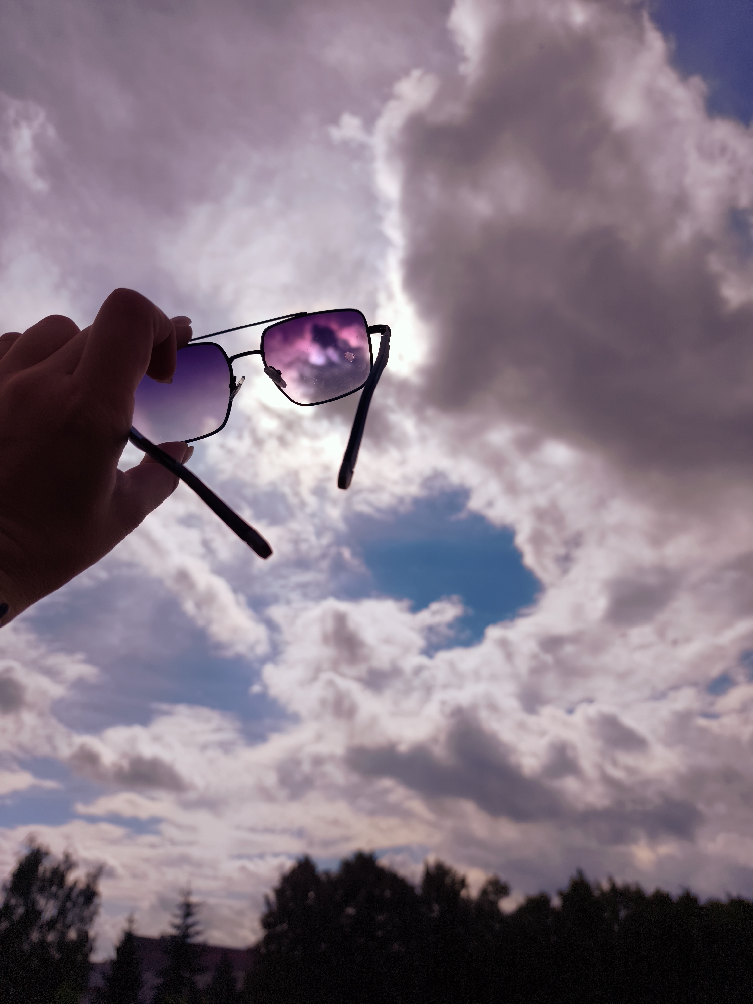 A hand holds up a pair of sunglasses against a cloudy sky with some trees visible in the distance. The sunglasses have black frames and gradient lenses that reflect the sky and clouds. The sky is filled with large, fluffy clouds with a pinkish hue, and there is a patch of blue sky visible. The trees at the bottom of the frame are dark and silhouetted. The overall scene appears to be taken outdoors, possibly in the late afternoon or early evening, given the lighting and the colors of the sky.