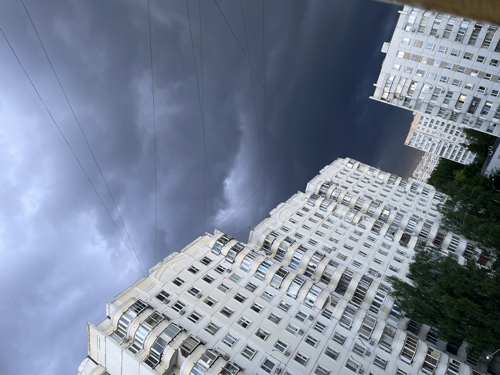 The image is a low-angle view of several white apartment buildings against a stormy sky. Power lines are visible against the sky. The buildings are densely packed, suggesting an urban environment. Green trees can be seen next to the buildings. The location is Moscow, Russia.