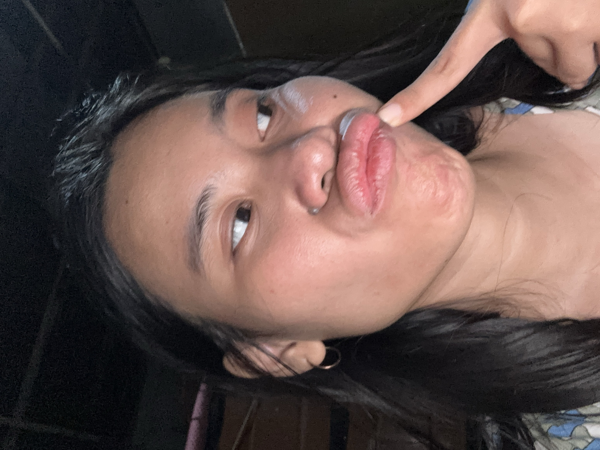 In the city of East Jakarta, Indonesia, a close-up, head-and-shoulders shot shows a young woman with dark hair and brown eyes making a puckered face. Her lips are pursed, and she is touching them with the index finger of her right hand. A white substance, possibly cream or makeup, is visible on her upper lip and cheek. She is wearing a small hoop earring in her left ear. The background is dark and appears to be indoors, with hints of texture suggesting a wall or ceiling.