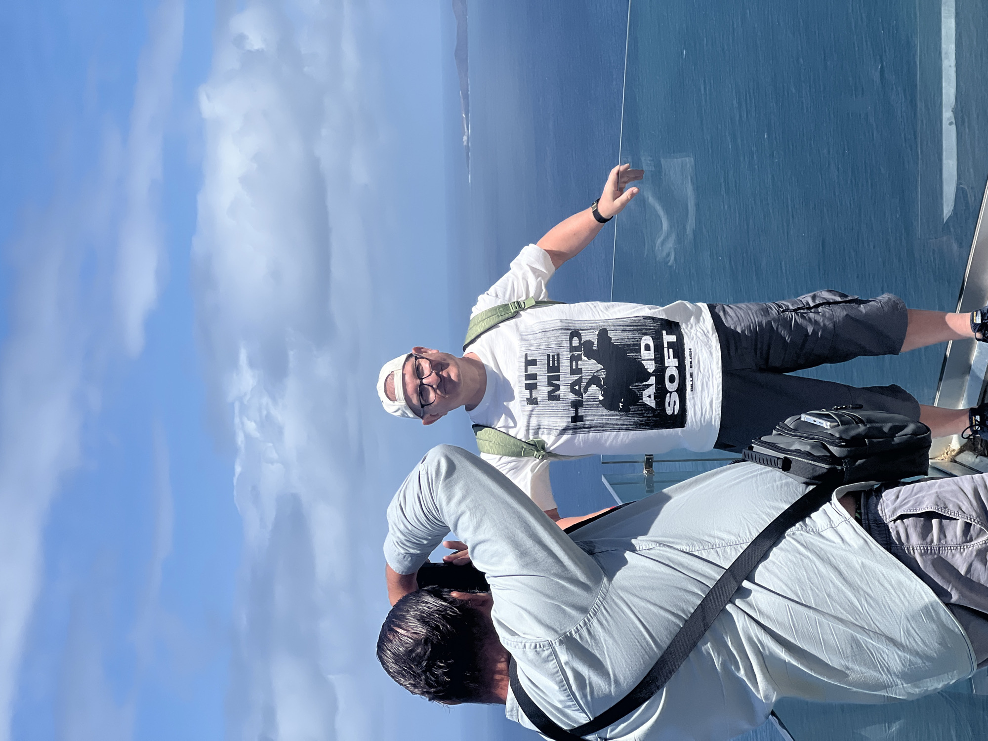 A medium shot captures two men on a glass observation deck. One man, wearing a white t-shirt with bold black text, grey shorts, and a white baseball cap, stands with one arm outstretched toward the blue sea visible beyond the deck. The other man, wearing a light grey shirt, is positioned slightly lower and is in the process of taking a photograph with a camera held up to his eye. A black camera bag is slung across his body. The background consists of a bright blue sky with scattered white clouds, and the expanse of the dark blue ocean. A land formation can be seen on the horizon. The photograph was taken in Barranco de Guayedra, Spain.