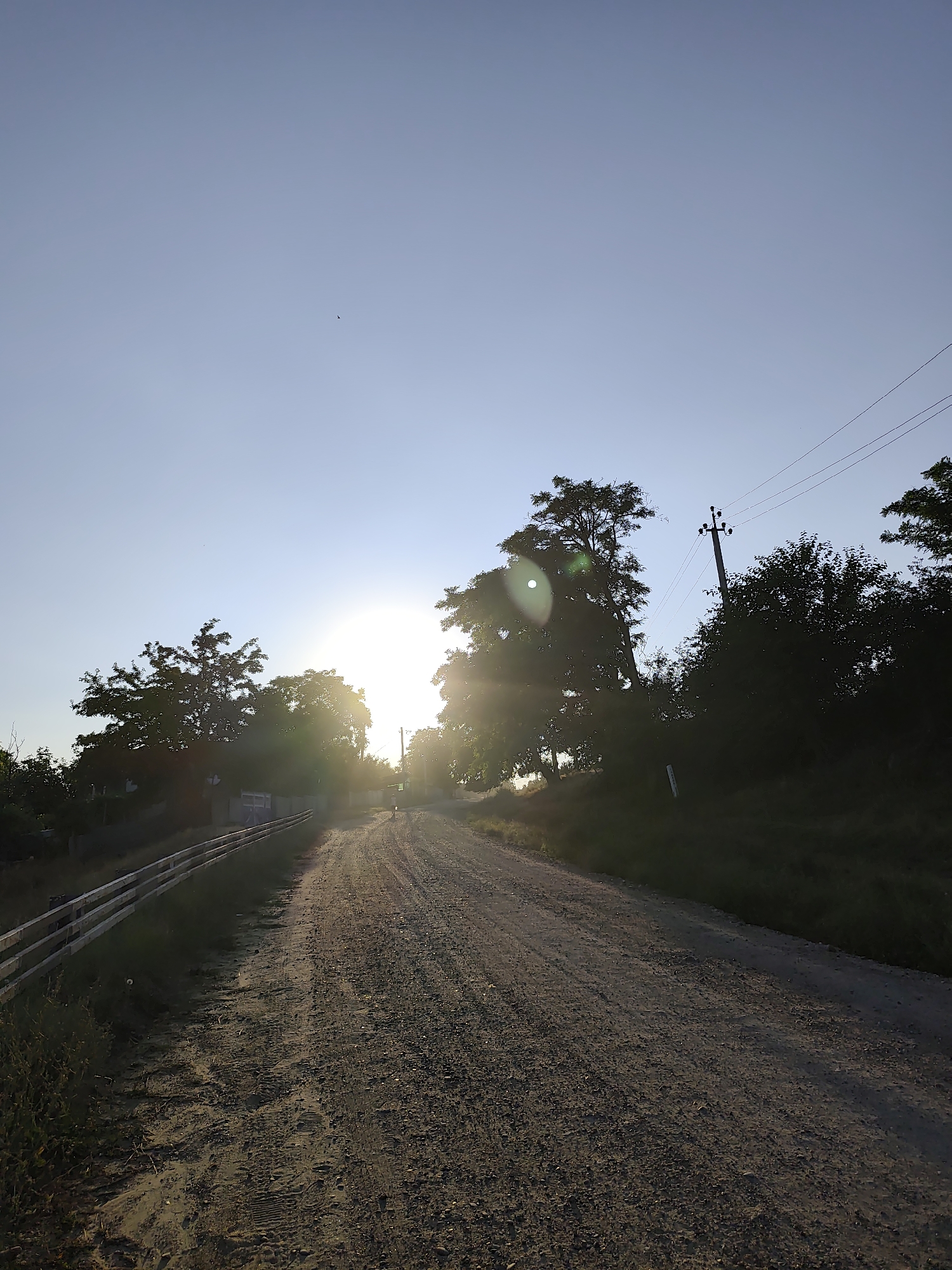 This is a full-shot, eye-level photograph taken outdoors on a sunny day. The scene depicts a gravel road leading into the distance, flanked by vegetation on either side. The sun is low in the sky, casting a warm glow and creating a lens flare. A silhouette of a person is visible in the distance on the road. Trees and power lines are present, with a wooden fence running alongside the left side of the road. The sky is a clear, light blue, with a small dark object, possibly a bird, visible in the upper left quadrant of the frame. The image appears to capture a rural or semi-rural setting, consistent with the village of Goian, Moldova.