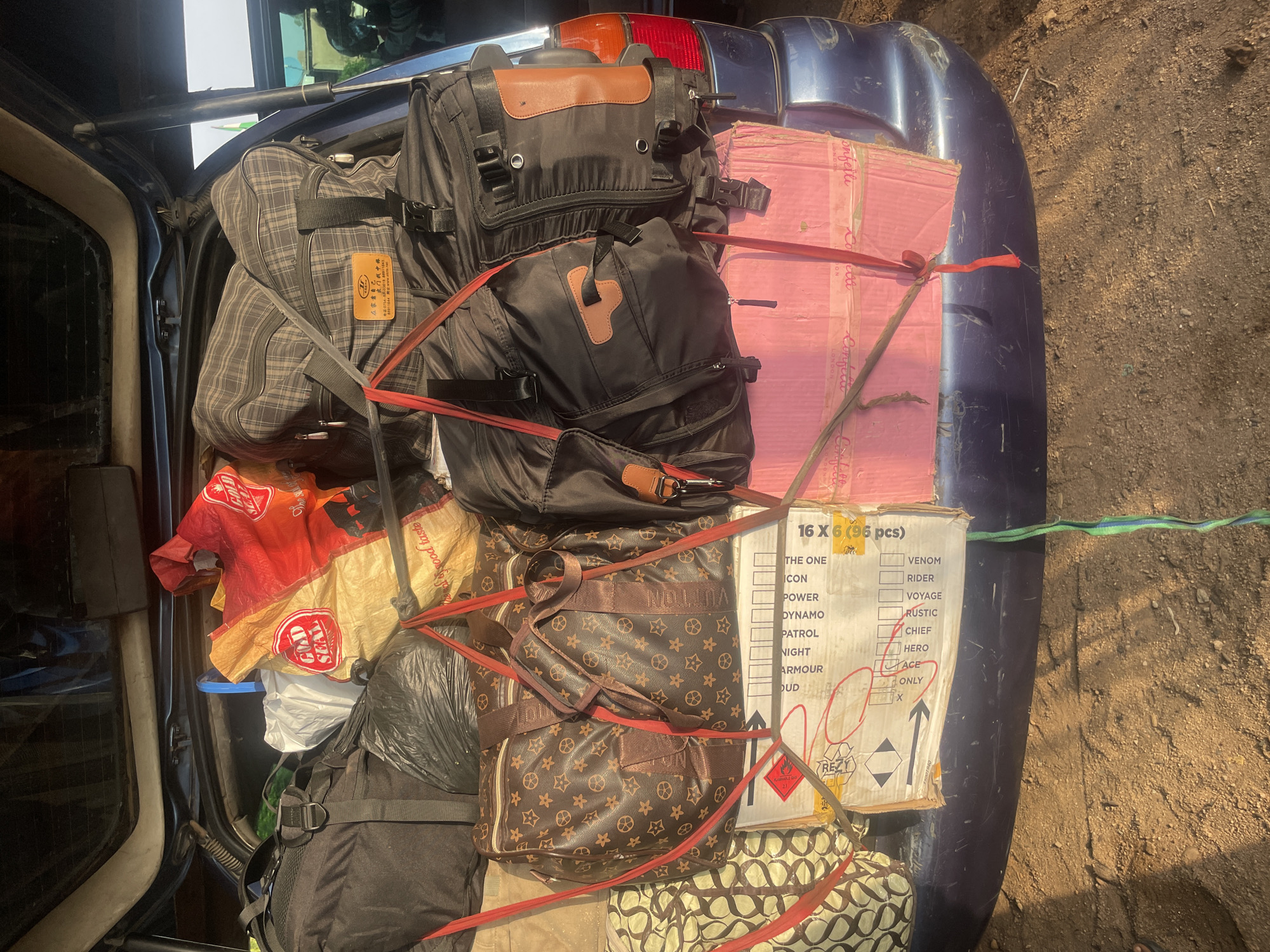 The image shows the back of a dark blue SUV loaded with luggage and boxes. The luggage includes several duffel bags, a large plaid suitcase, and a patterned duffel bag with a Louis Vuitton-inspired print. There are also several cardboard boxes stacked in the rear. One box is pink and another has a label with text including "16 X 6 (96 pcs)" and several other words that appear to be product names. Red and green straps are used to secure the contents. The car is parked on a dirt road in what appears to be an outdoor area, with dry grass and dirt visible on the ground. The scene is consistent with a vehicle being prepared for or having just completed a journey in Wauro Jabbe, Nigeria, a town known for its agricultural activities and rustic environment.