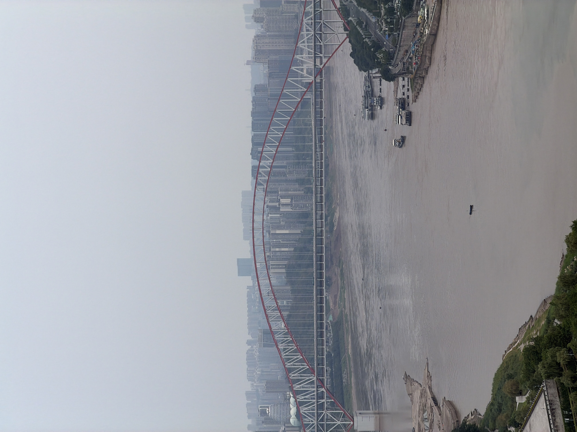 This is an aerial, full-frame, landscape photograph of the Yangtze River and the Chongqing Yangtze River Bridge, viewed from Yuzhong District, China. The sky is overcast with a light haze, obscuring the distant city skyline, which is densely packed with numerous high-rise buildings. The bridge, a prominent steel arch structure, spans the wide, muddy-brown river. The bridge features a red arch and white supports, with a lower level roadway carrying traffic. Several boats and barges are visible on the river, including larger vessels docked along the right bank and smaller boats in the water. The riverbanks are lined with trees and vegetation, and the right bank shows a developed embankment with roads and lighting. The overall impression is an urban industrial landscape characterized by large infrastructure and a hazy atmosphere.