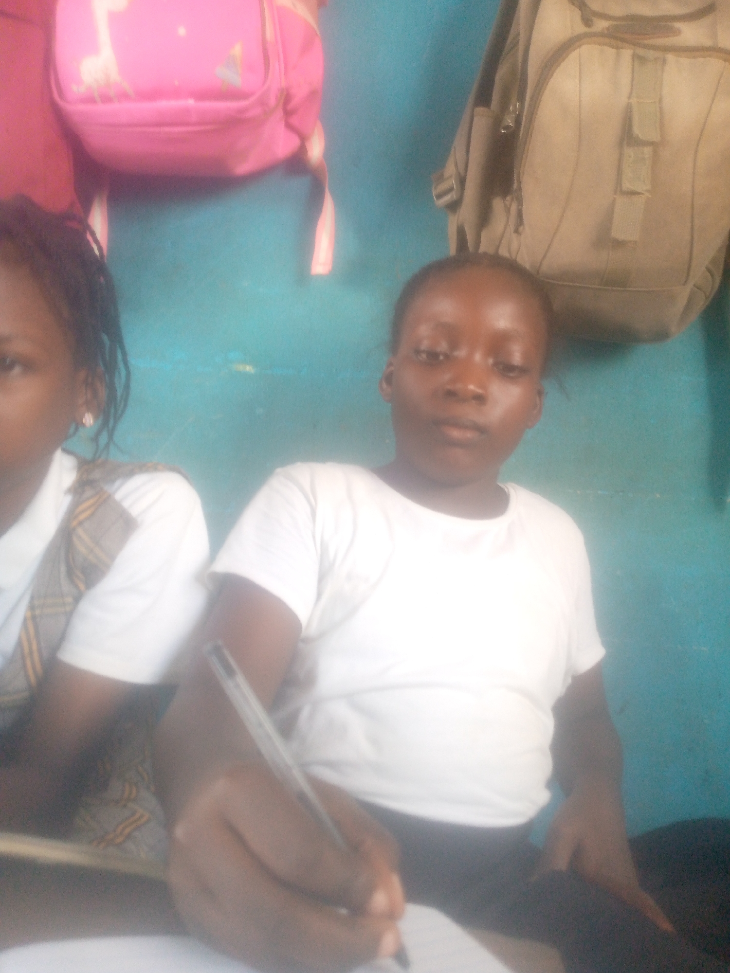 Two young girls are seated at a desk in what appears to be a classroom in Unknown, Unknown. The girl on the right is writing with a pen on a piece of paper. Both girls are wearing school uniforms. Behind them on the wall hang a pink backpack and a tan backpack. The wall is blue.