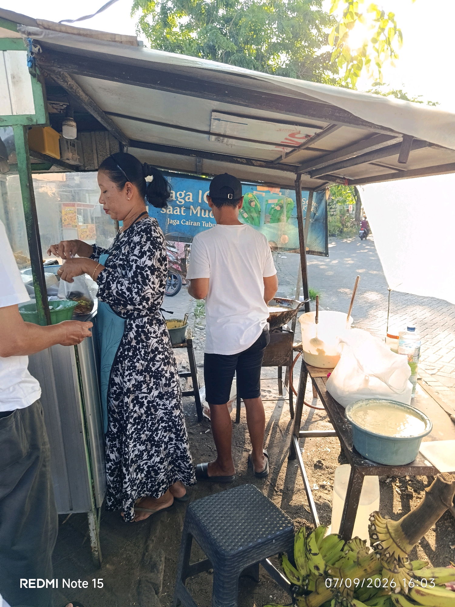 An outdoor food stall, likely in Keboansikep, Indonesia, is depicted. The stall has a white tarp roof supported by metal poles. A woman in a black and white floral dress and a blue apron is seen on the left, preparing food. She is holding a plastic bag and appears to be serving something. To her right, a man in a white t-shirt and black shorts stands facing away from the camera, looking at the stall. In front of the stall, a table holds various items including a large bucket of pale yellow liquid, a white plastic bag, and a bottle of water. There are also bunches of green bananas on the ground. The ground is dirt and gravel. Sunlight is visible filtering through trees in the background. The image has a timestamp at the bottom right indicating "07/09/2026 16:03" and "REDMI Note 15" at the bottom left.