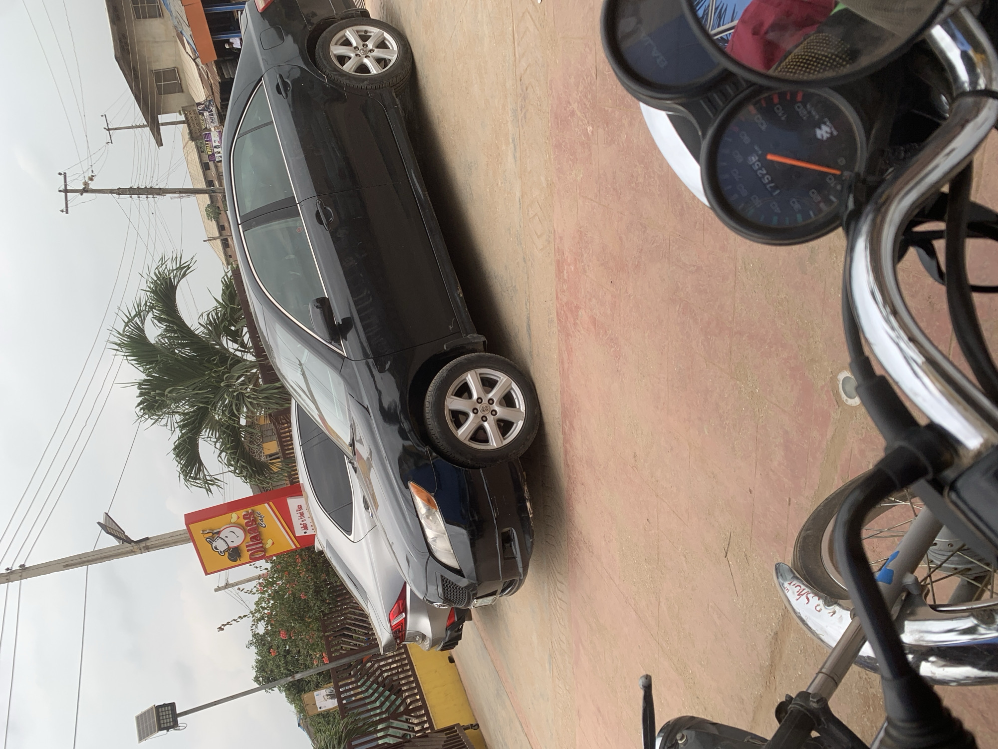 The image depicts a street scene in Shagamu, Nigeria. In the foreground, the handlebars and dashboard of a motorcycle are visible, with a speedometer showing "175258". Behind the motorcycle, two cars are parked on a paved surface. The car on the right is a black sedan, and the car on the left is a silver sedan. A sign for "Ollaass Cafe" with a cartoon cow logo is visible in the background, along with palm trees and buildings. Overhead, power lines stretch across the sky, and a street light with a solar panel is mounted on a pole. The overall environment suggests a commercial or roadside area.