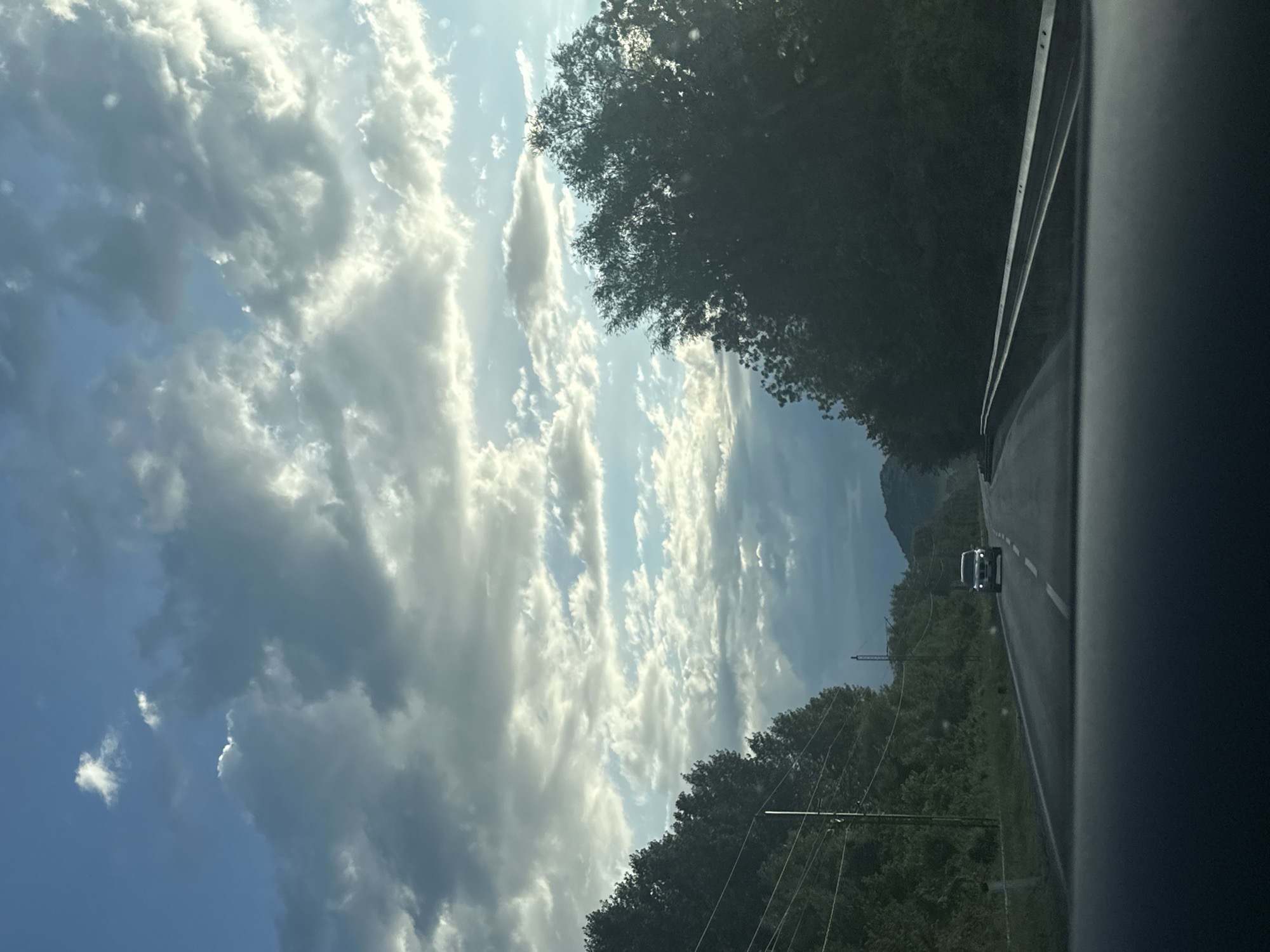 The image presents a daytime view from inside a moving vehicle, looking forward along an asphalt road in a natural, mountainous setting near Bistrice, Montenegro. The sky dominates the upper portion of the frame, showcasing a mix of deep blue sky and bright, voluminous white cumulus clouds, illuminated by strong sunlight, suggesting a sunny day.
The road, with a visible white shoulder line and a partial dashed center line, extends into the distance. Lush, dense green trees and vegetation line both sides of the road, forming a thick forest that appears to cover rolling hills or the base of mountains. In the far distance, on the left side of the road, outlines of mountains are discernible under the cloudy sky.
Further down the road, a white car, possibly an SUV or van, is visible in the same lane, with its headlights on. Utility poles and power lines are also evident through the trees on the left side of the road, indicating human infrastructure in the natural landscape. The right edge of the vehicle's interior (likely the dashboard or windshield frame) occupies the foreground on the right side of the image. No visible text is present. The scene captures a moment of travel through a scenic, rural or semi-rural mountainous region.