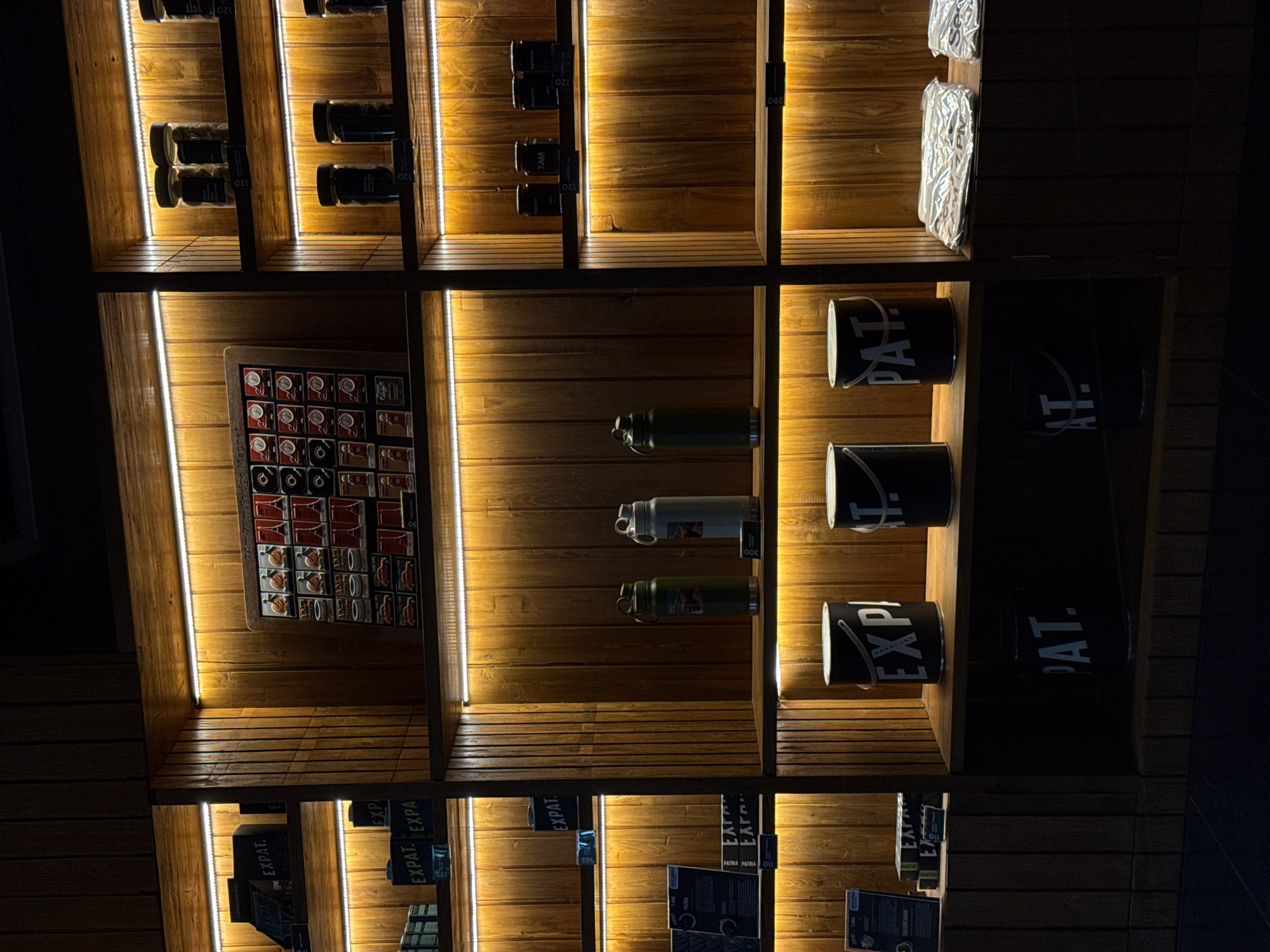 Captured at night, the image showcases a brightly lit, wooden shelf display inside a store, likely in the Ubud District, Indonesia. The shelves are divided into compartments holding a variety of merchandise, including sealed packages, water bottles, and cans labeled "EXPAT" and "PATRIA." A framed display of magnets is also present. The artificial lighting highlights the wooden texture of the shelves, casting a warm glow on the products.