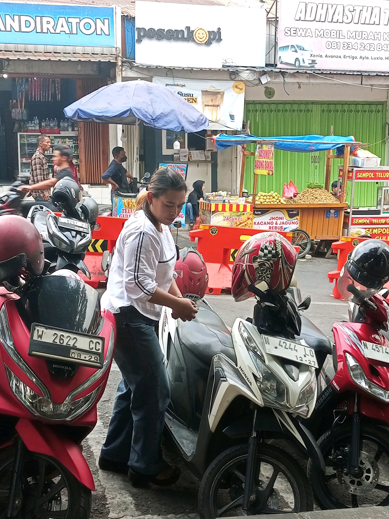 The image depicts a street scene in Batu, Indonesia, characterized by a row of food stalls and parked motorcycles. In the foreground, several motorcycles are parked, some of which have helmets placed on them. A woman in a white shirt and blue jeans is standing next to one of the motorcycles, looking down. Behind her, more motorcycles and a blue umbrella shade are visible.
The food stalls display various food items, including what appear to be snacks and drinks. Signs above the stalls advertise "SUSU," "JASUKE," "JHONI KACANG & JAGUNG REBUS," and "SEMPOL TELUR." The background shows buildings with signage, including "ANDIRATON" and "ADHIASTHA THY," suggesting commercial activity. The overall environment appears to be an outdoor marketplace or busy street.