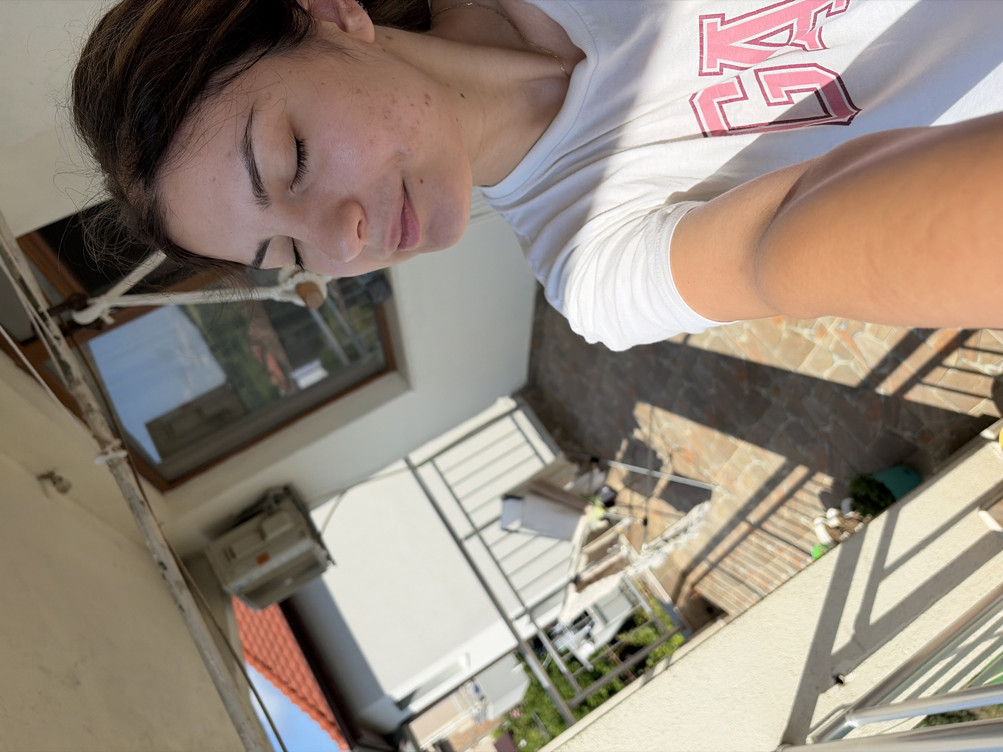 A young woman is taking a selfie on a balcony in Tsarevo, Bulgaria, with her eyes closed and a slight smile. She is wearing a white t-shirt with pink lettering. The balcony overlooks a residential area with houses and a clear blue sky. The sun is shining, casting long shadows across the tiled balcony floor. An air conditioning unit is visible on the exterior wall of the building. The woman's skin shows some imperfections, including acne marks and freckles. The overall scene depicts a relaxed moment outdoors on a sunny day.