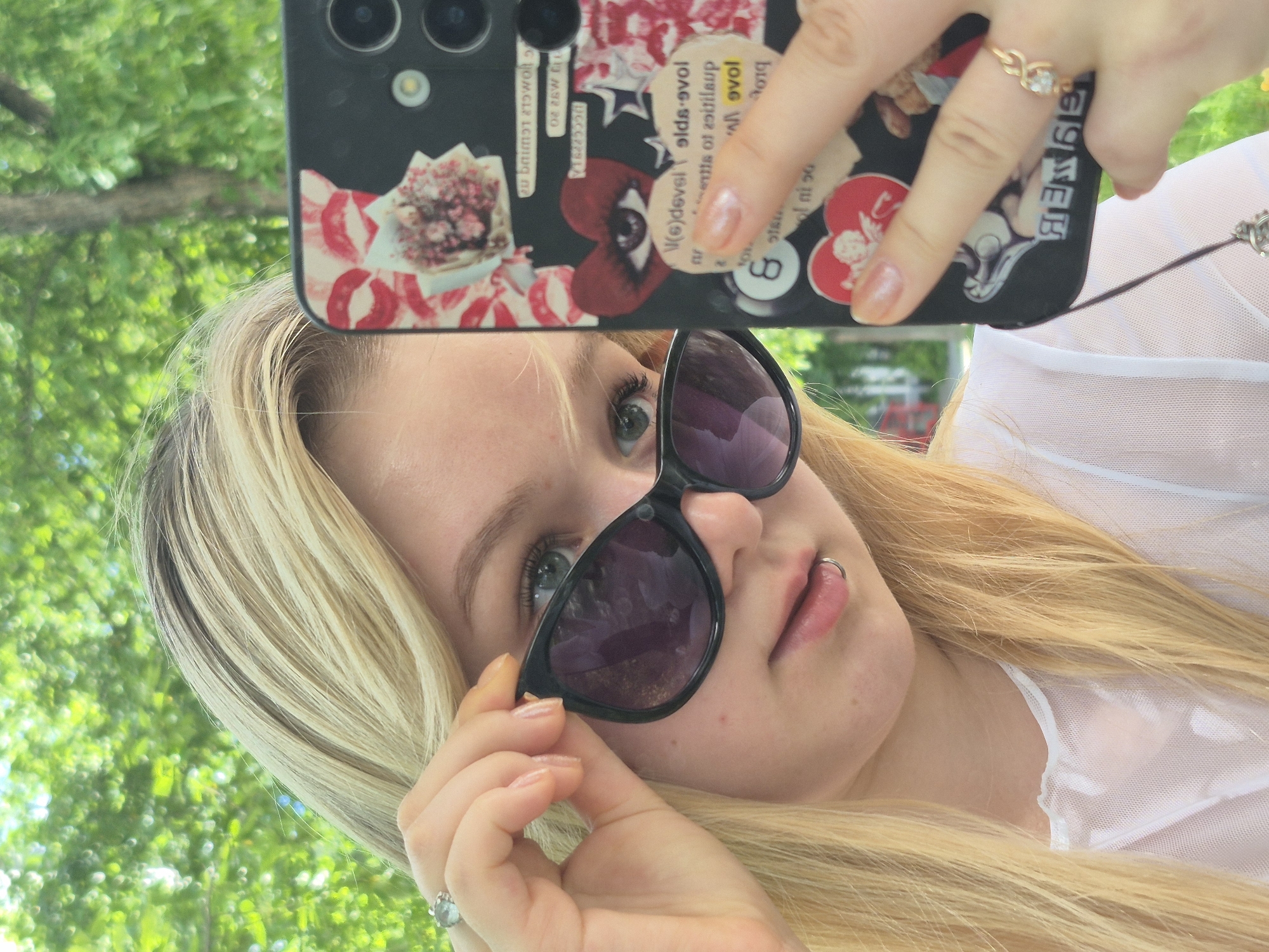 A young woman with blonde hair and sunglasses is taking a selfie outdoors. She is holding her sunglasses with her left hand, revealing one eye. Her right hand is holding her phone, which has a black case decorated with stickers. The background is out of focus and appears to be greenery, suggesting she is in a park or garden. The image does not contain any information that allows for its localization in Chaikovsky, Russia.