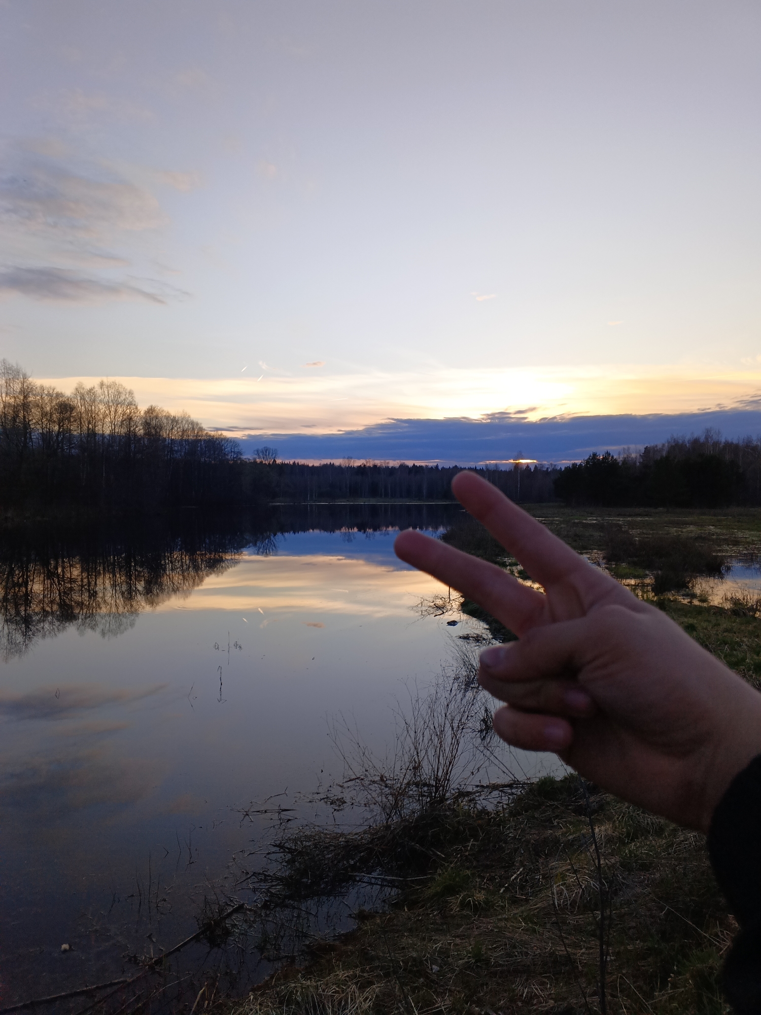 The image shows a landscape at sunset with a body of water in the foreground, likely a lake or wide river. Trees line the shore, reflected in the water. The sky is a gradient of colors, ranging from pale blues and purples to orange near the horizon. A person's hand, making a peace sign, is partially visible in the foreground on the right. The surrounding area appears to be a natural environment with vegetation and grass. Based on the location provided, Красный Маяк, Russia, it can be inferred that the landscape is typical of the Russian countryside.