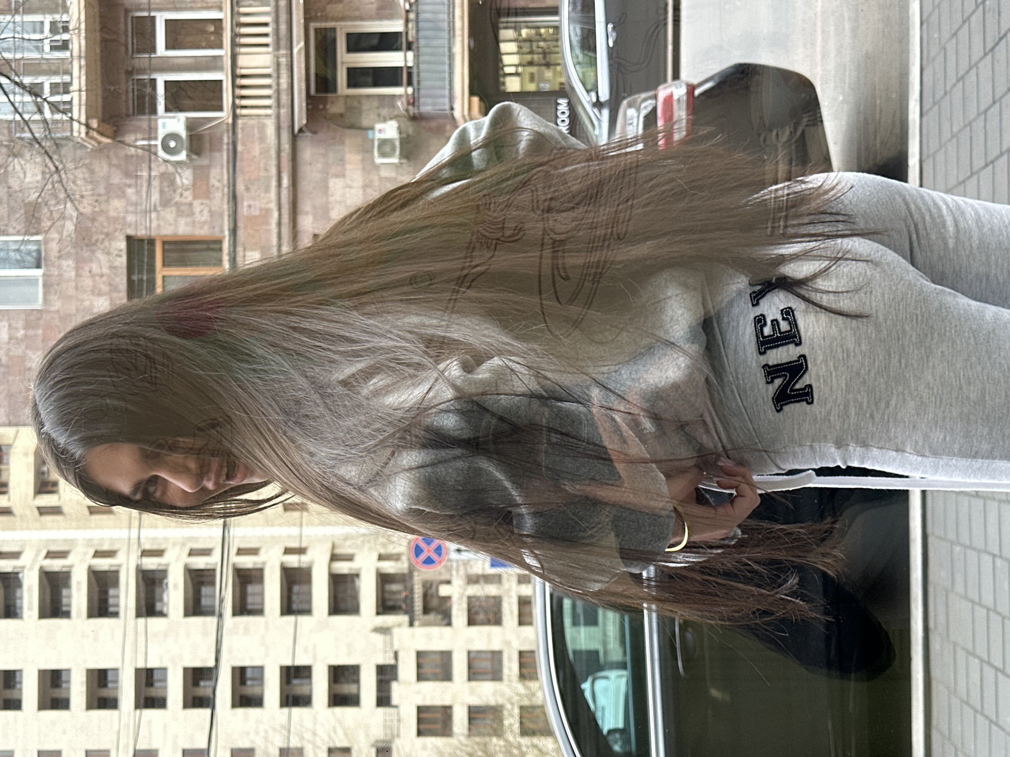 The image displays a female individual, primarily seen in reflection against a glass surface, standing on a light grey tiled sidewalk. She is observed from a slightly rear-right perspective, turning her head over her right shoulder, with long, straight, dark brown hair extending past her waist, partially obscuring her face. Her expression indicates a slight smile. She wears a grey hooded sweatshirt and matching grey sweatpants, featuring the embroidered text "NEX" in dark letters on the left posterior. A thin, gold-toned bracelet is visible on her right wrist, with her right hand positioned near the drawstring of her sweatpants.
The background, visible through the reflection, presents an urban street scene consistent with the city of Yerevan, Armenia. To the left, a modern, multi-story building constructed of light-colored stone features numerous recessed balconies and windows. To the right, an older building, characteristic of Yerevan's architecture, is constructed of reddish-brown tuff stone. This older structure includes a decorative balcony with balusters on the second floor and an external air conditioning unit mounted below a window. On the adjacent street, a dark-colored sedan is visible. A blue circular sign with a red 'X' (indicating a prohibitory traffic regulation) is also discernible in the reflection. Bare tree branches are visible against the older building, suggesting a non-foliage season.