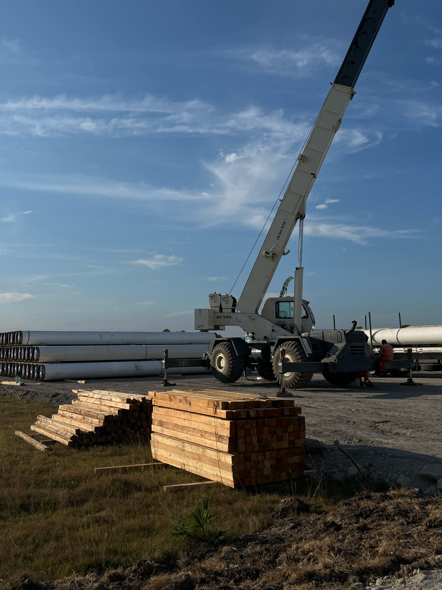 A construction site scene unfolds under a blue sky with scattered clouds. In the foreground, stacks of light-colored lumber are neatly arranged on grassy ground. To the left, a larger, angled stack of lumber rests on the grass. Beyond the lumber, a row of large, white cylindrical pipes is stacked horizontally. Dominating the midground is a large, white mobile crane with its boom extended upwards. The crane's cab and wheels are visible. A person in an orange shirt is seated on a trailer to the right of the crane. The ground is a mix of dirt and gravel. The scene appears to be an outdoor storage or staging area for construction materials.