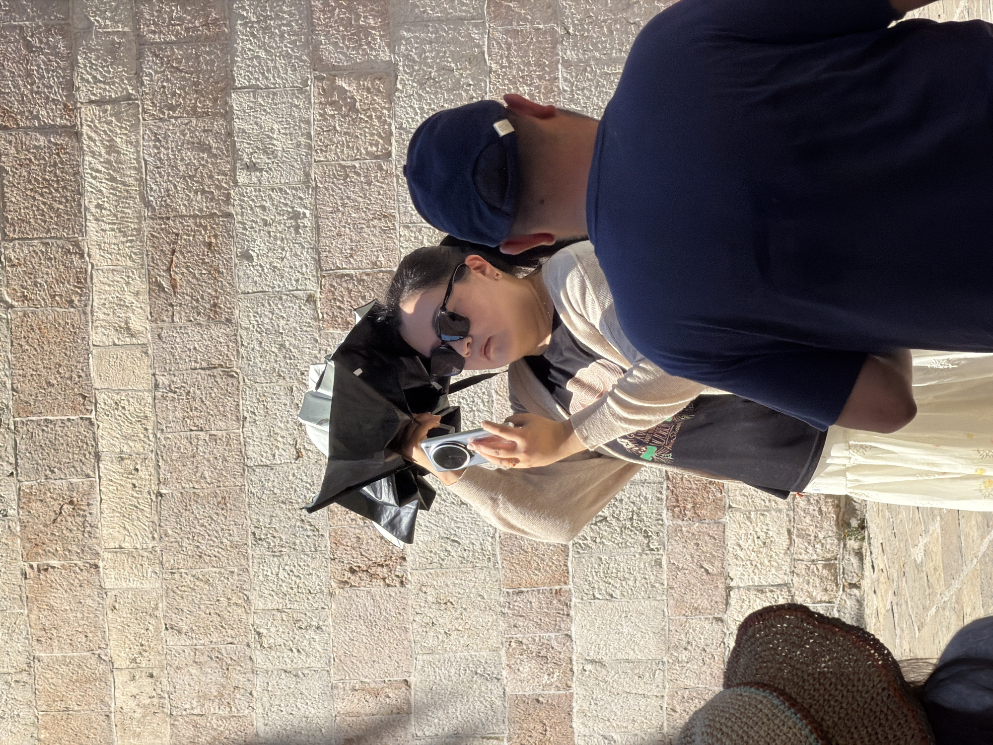 In Budva, Montenegro, a woman with dark hair and sunglasses holds a small black umbrella over her head and a silver digital camera, framing a shot against a stone wall. She wears a light-colored cardigan over a dark t-shirt and a long, flowing cream skirt. To her right, the back of a man wearing a navy baseball cap and a dark blue t-shirt is visible, facing away from the camera. In the lower left of the frame, the straw hat and shoulder of another person are partially visible. The background is a textured wall constructed from rectangular light-colored stones. The ground is paved with cobblestones.