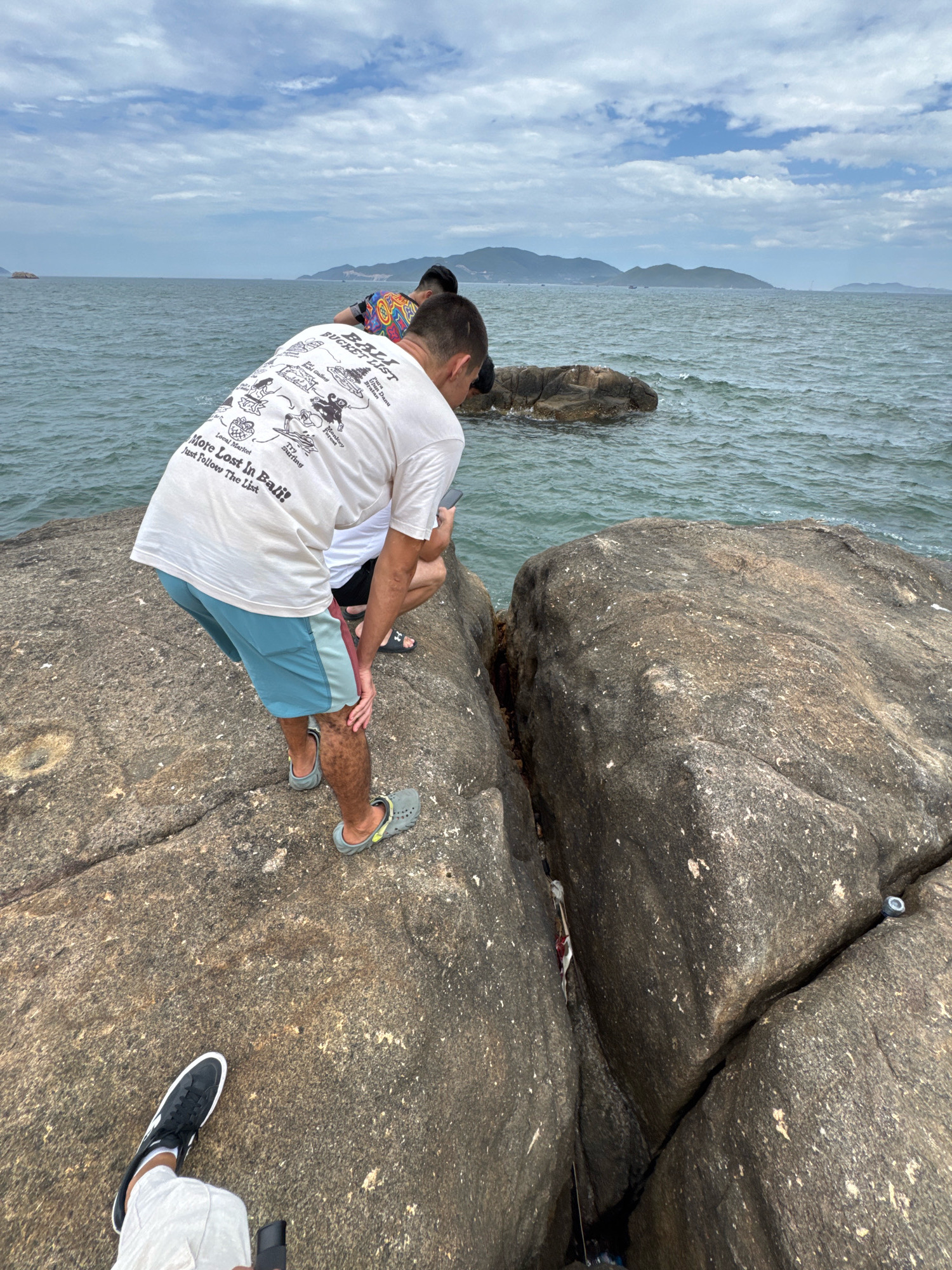 The image depicts three individuals near the rocky shoreline of the South China Sea, likely in Nha Trang, Vietnam, given the typical coastal scenery. In the foreground, a person wearing white pants and black sneakers is holding a camera, indicating they are taking the photo or recording. Behind them, a person in a white t-shirt with a graphic print and blue shorts is bending over, looking into a crevice between two large rocks. Their t-shirt features text that appears to say "More Lost in Bali! Just Follow The List" and various drawings. A third individual, wearing a brightly colored patterned shirt, is partially visible in the background, also looking towards the sea. The environment consists of large, weathered granite boulders forming a natural barrier against the calm, grey-blue ocean. Distant landmasses, possibly islands or the mainland coast, are visible on the horizon under a cloudy sky. The scene captures a moment of exploration or observation of the coastal landscape.