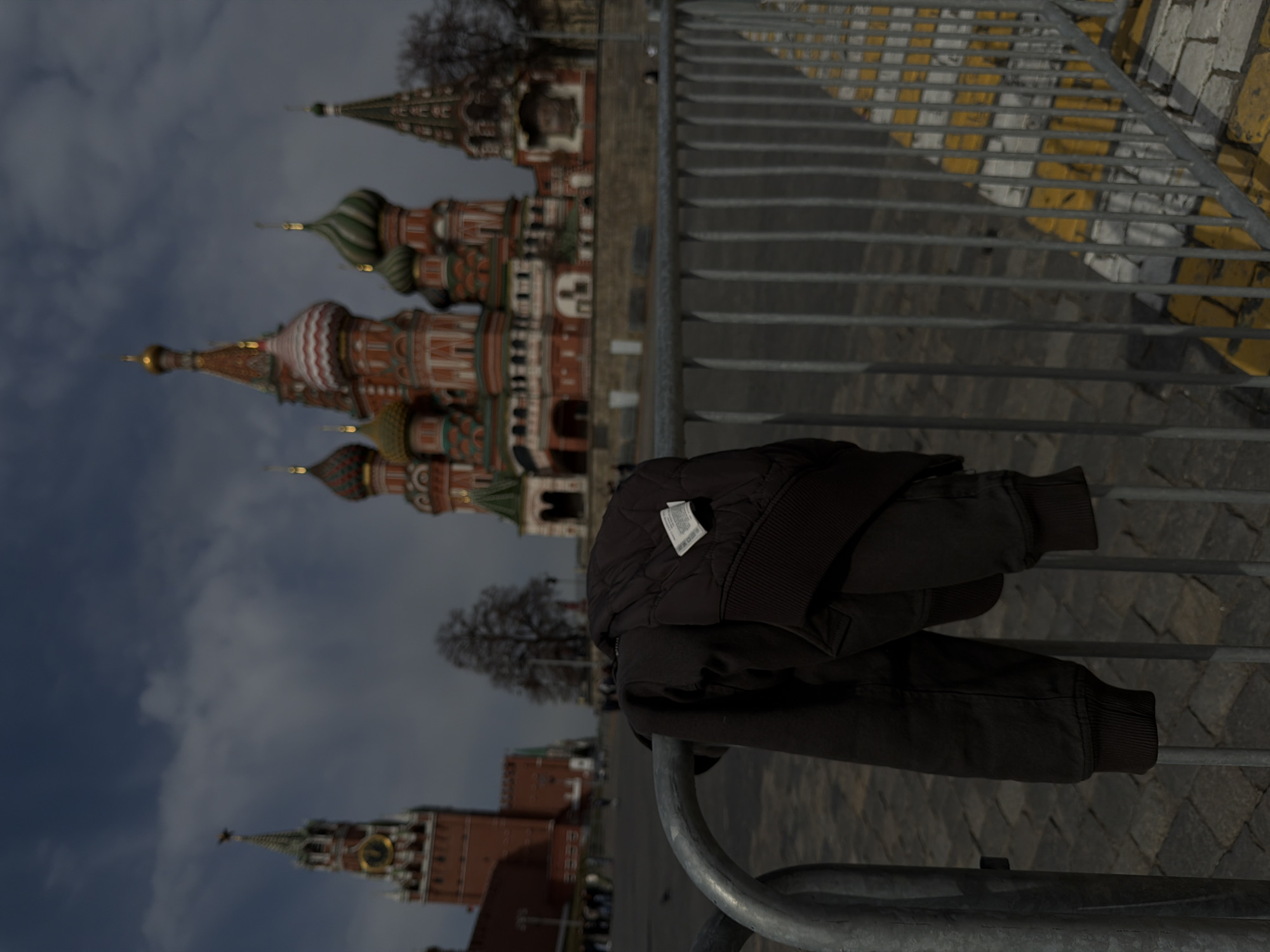 A jacket hangs on a metal barrier in Moscow, Russia. The blurred background reveals Saint Basil's Cathedral and the Spasskaya Tower of the Kremlin. The sky is overcast. The location is near the cobblestone plaza.