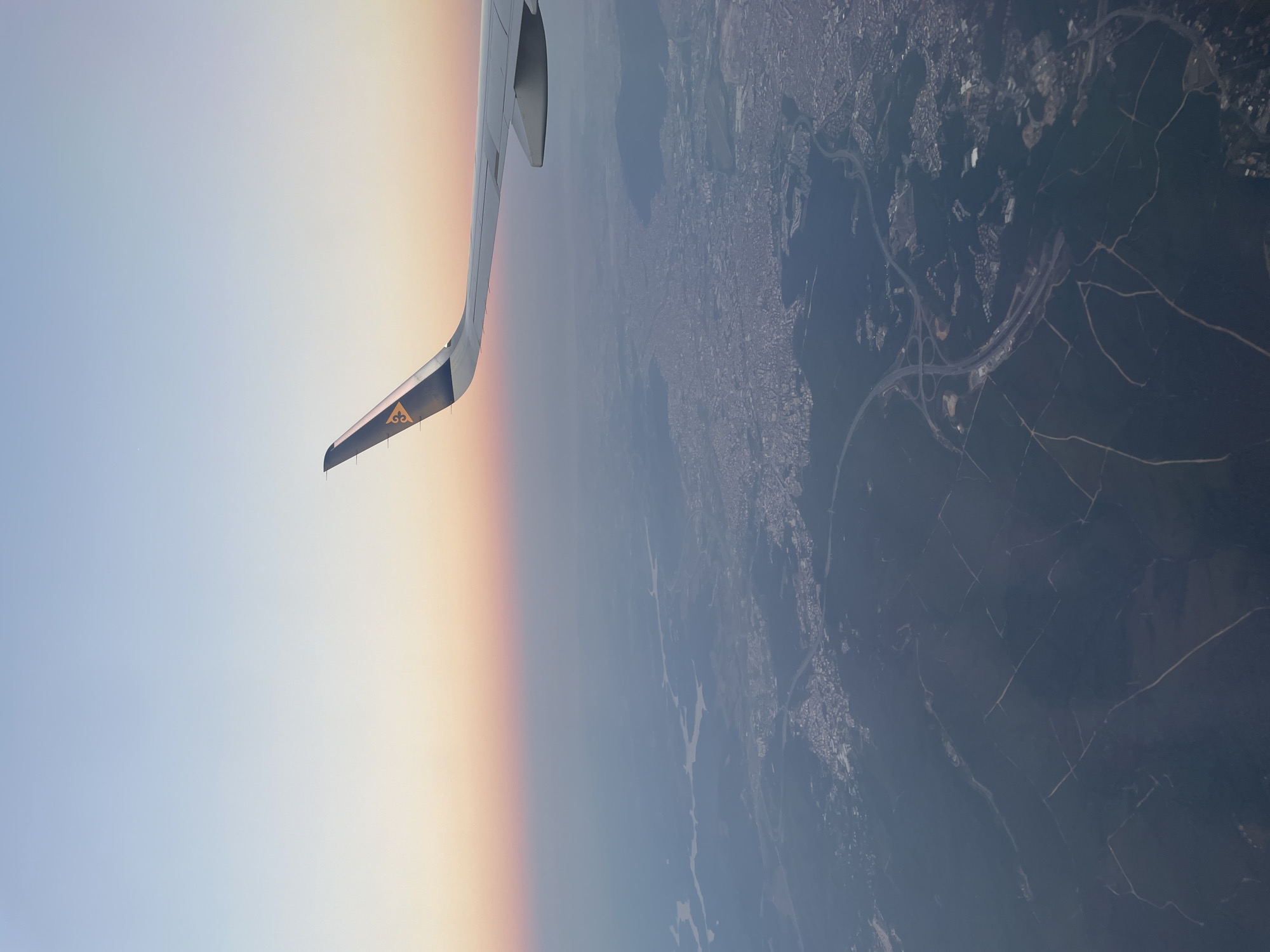 This is an aerial photograph taken from an airplane, capturing a sunset over the city of Beykoz, Turkey. The airplane's wing, featuring the logo of a stylized bird in gold on a dark blue background, is visible in the upper right portion of the frame. Below the wing, the sky transitions from a pale blue at the top to soft hues of pink and orange near the horizon, indicating the setting sun.
The landscape below is a mix of urban development and natural terrain. A large city sprawls across the middle ground, characterized by dense clusters of buildings and streets. Further in the distance, a body of water, likely the Bosphorus or a related waterway, is visible, with winding rivers or channels cutting through the landscape. The foreground is dominated by dark green, hilly terrain, crisscrossed by irregular patterns of lighter colored lines, which appear to be roads or pathways. The overall atmosphere is serene, with the soft light of the sunset casting a gentle glow over the scene.
