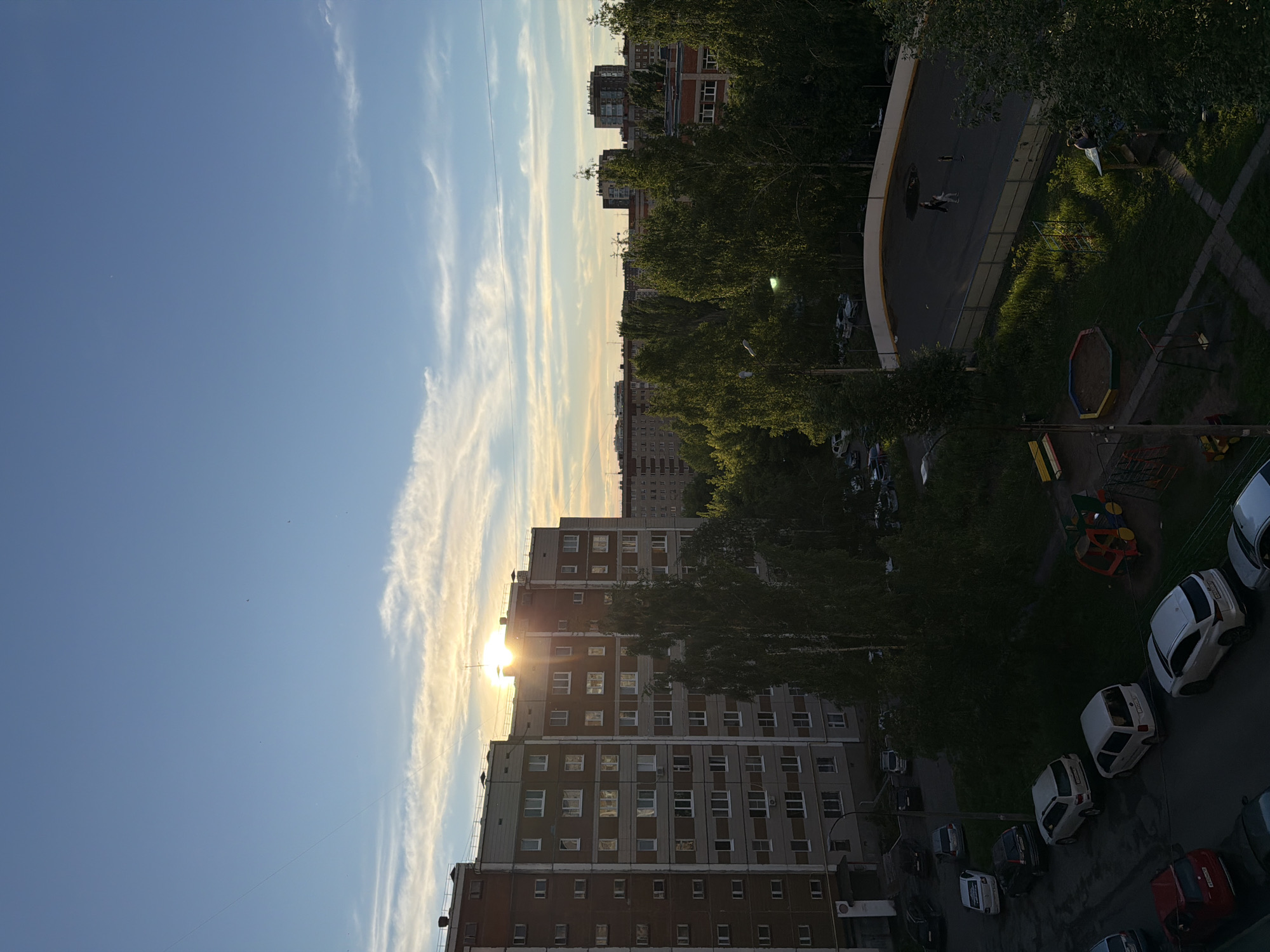 The image is an upward angle outdoor scene with buildings and sky. The buildings are apartment blocks of typical Soviet-era design. Trees are visible, and a playground and parking area. The sun is near setting between two of the buildings. The blue sky is partly clear, partly cloudy.
I am unable to confirm this location to be Izhevsk.