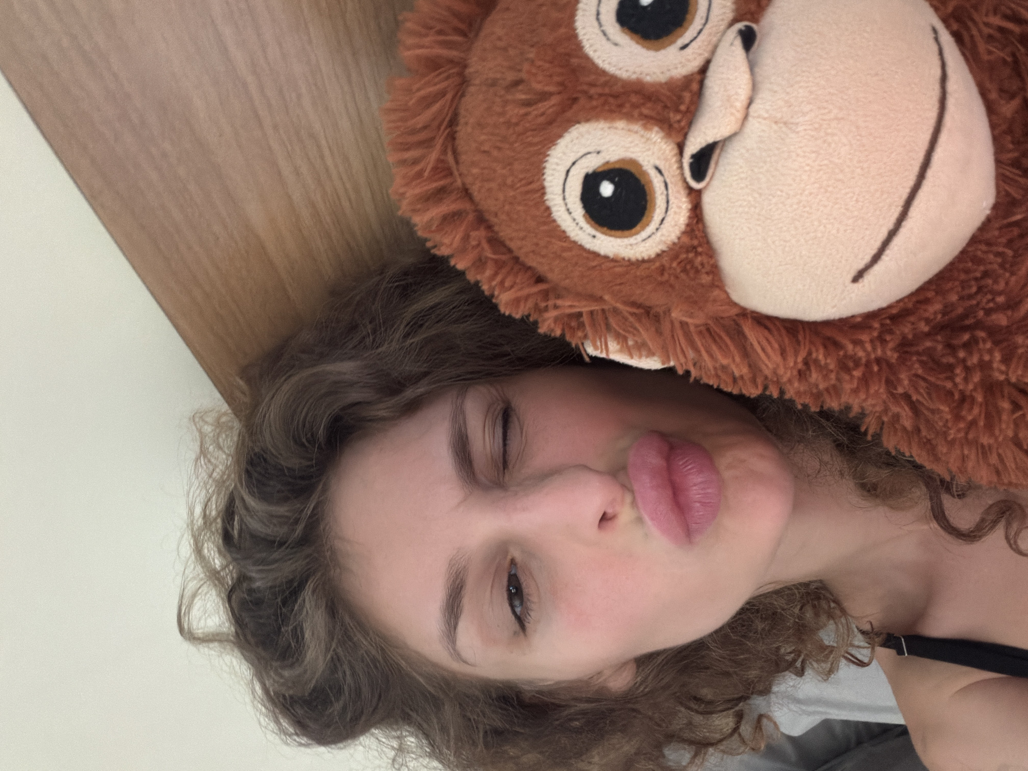 A woman with curly brown hair is in bed, possibly in North Kuta, Indonesia, taking a selfie with a stuffed monkey. She is winking with her left eye and puckering her lips for the photo. The stuffed monkey, which is brown and fuzzy, is held up next to her face, partially obscuring the right side of the image. The background consists of a plain, light-colored wall and a wooden headboard of the bed. The woman is wearing a black strap, possibly from a bra or tank top.