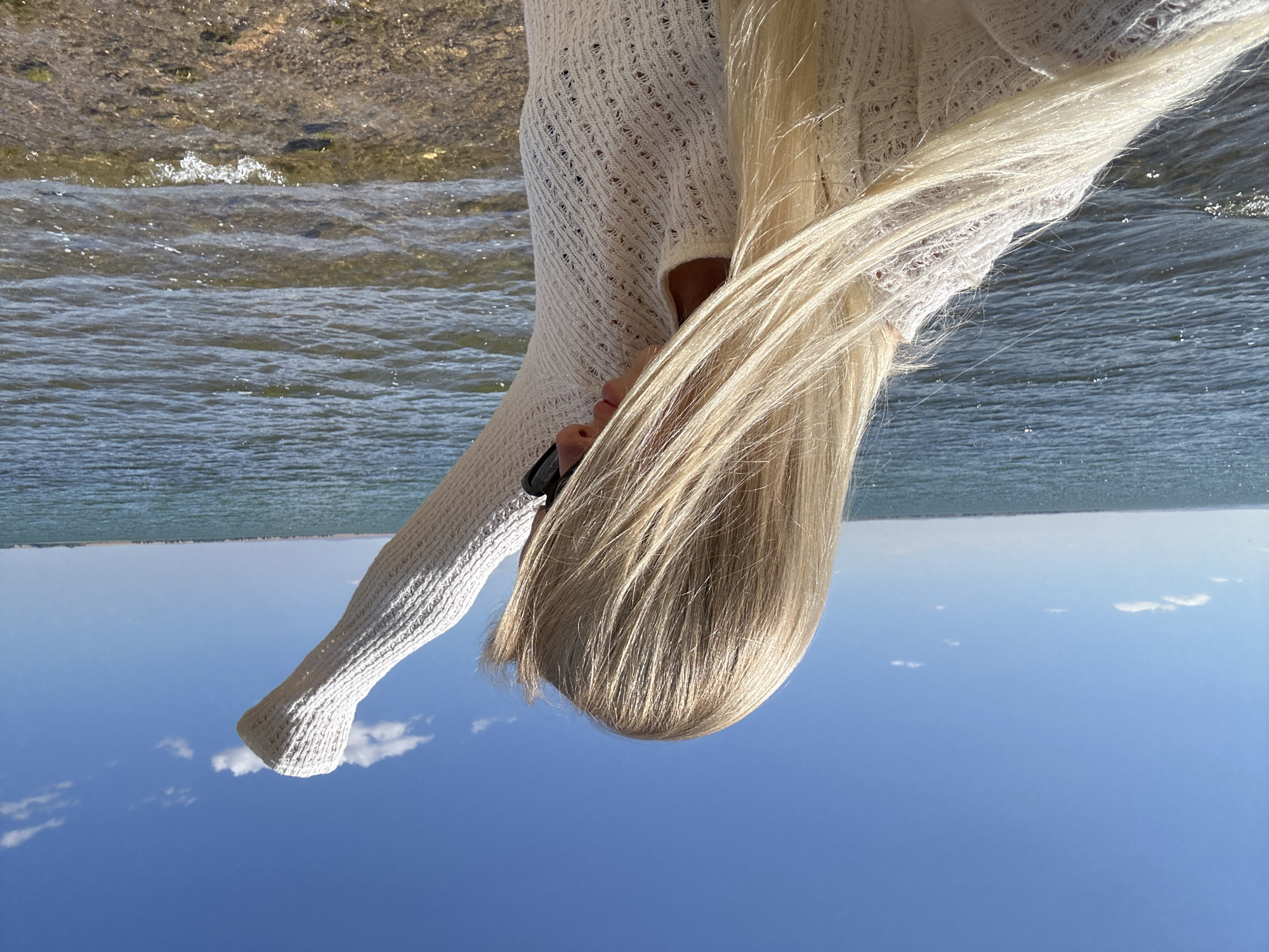 A medium shot captures a person with long, blonde hair standing by the water's edge. The person is facing away from the camera, with their right arm raised in the air. They are wearing a white, textured, long-sleeved top. The background consists of a calm body of water under a clear blue sky with a few scattered clouds. The shoreline is visible in the foreground, composed of small rocks and pebbles. The location is the city of Gornyi Erik, Russia.