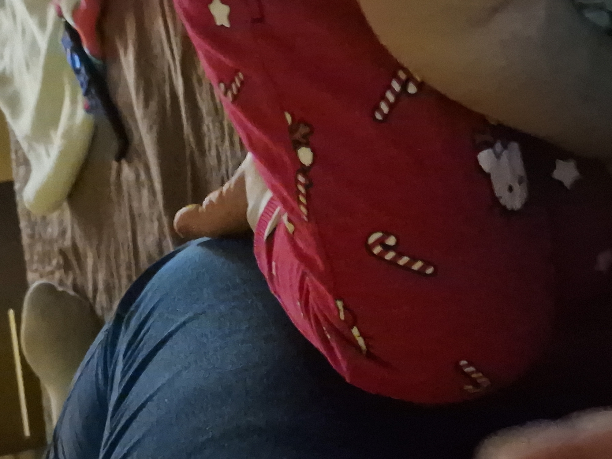 The image shows a person wearing blue jeans and red patterned pajama pants with candy cane, reindeer, and star designs. The person is resting their bare foot on their leg, and there is bedding visible in the background. The image provides no information regarding location in Byala Slatina, Bulgaria.