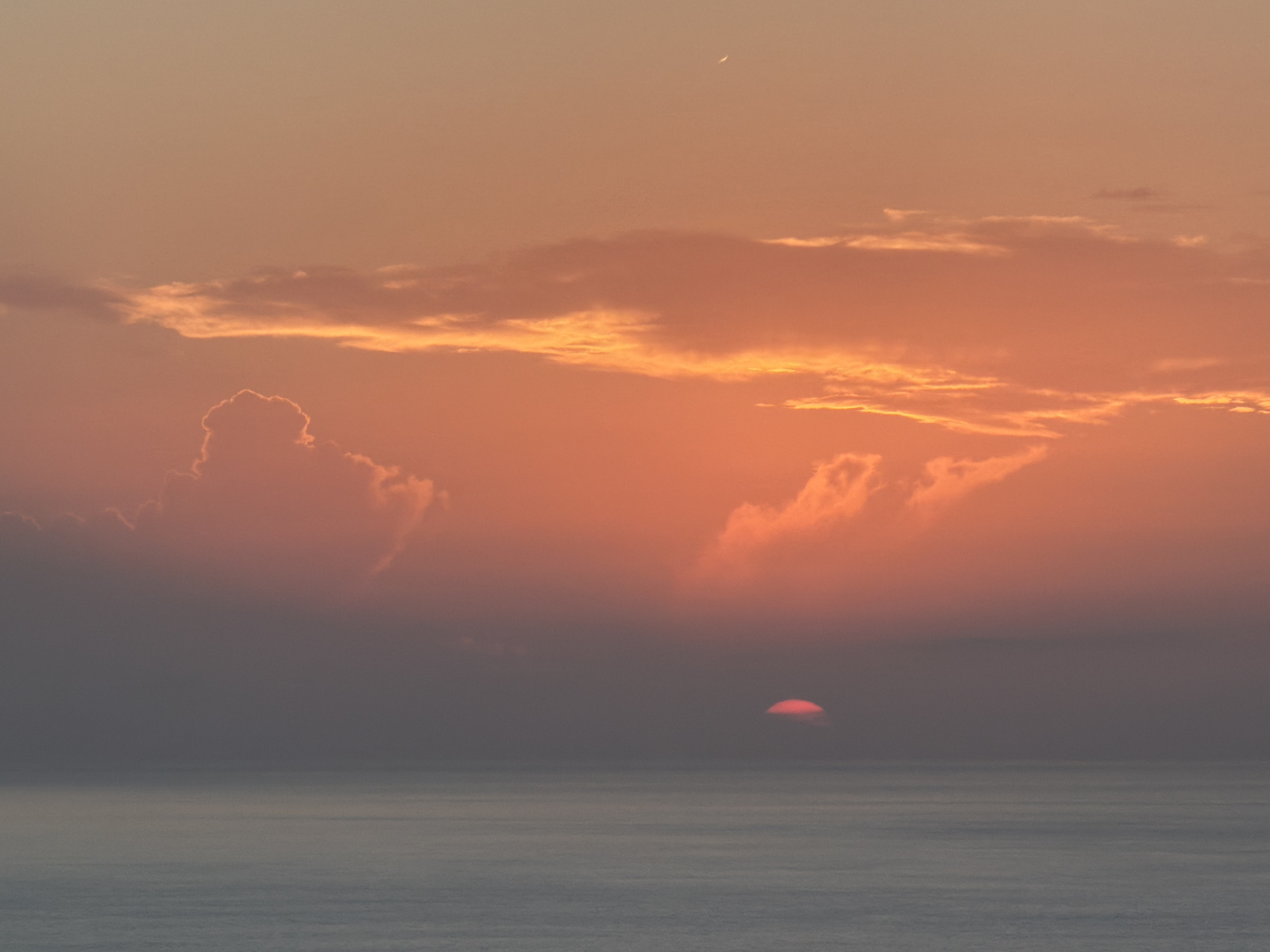The image captures a serene sunset over the Mediterranean Sea, viewed from Sant Antoni de Portmany, Spain. The sky is awash in hues of orange and pink, with scattered clouds catching the vibrant light. The sun, a small crimson orb, is partially submerged below the horizon, casting a soft glow on the water's surface. The sea itself appears calm, with gentle ripples reflecting the twilight colors. There are no people or man-made structures visible, emphasizing the natural beauty of the coastal landscape. A faint crescent moon is visible in the upper portion of the sky.