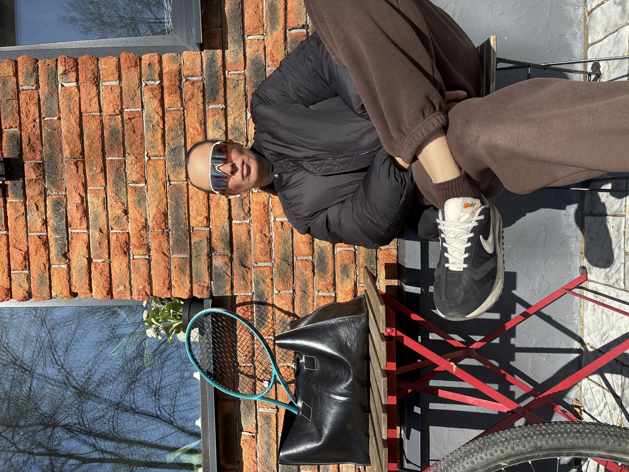 A person with sunglasses and a black jacket and brown sweatpants is seated in front of a brick building in Chaikovsky, Russia. The individual has one leg crossed over the other. They are wearing black and white sneakers. A black leather bag and a tennis racket are next to them. A bicycle wheel is visible on the ground. The reflection of trees can be seen in a window of the building. The scene is well-lit with sunlight. The area is paved with gray tiles. A red folding chair is also visible.